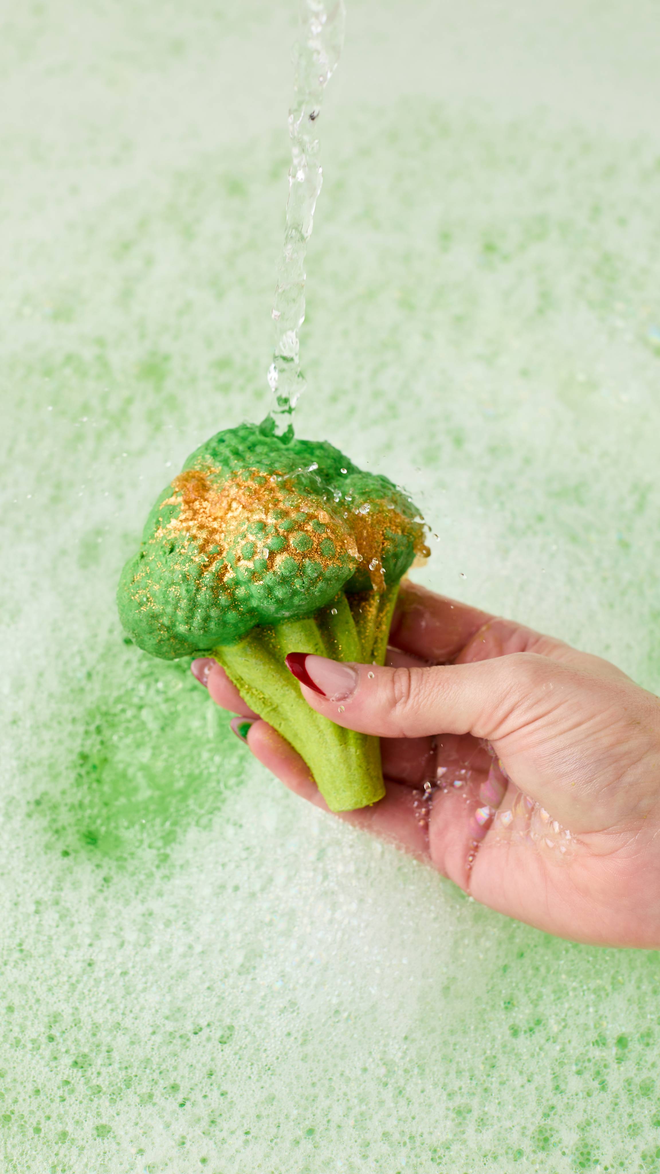 The image shows a hand with long nails holding the vibrant green Sparkly Broccoli bubble bar under a stream of water that splashes off. Delicate green water sits below, covered in a blanket of thick bubbles. 