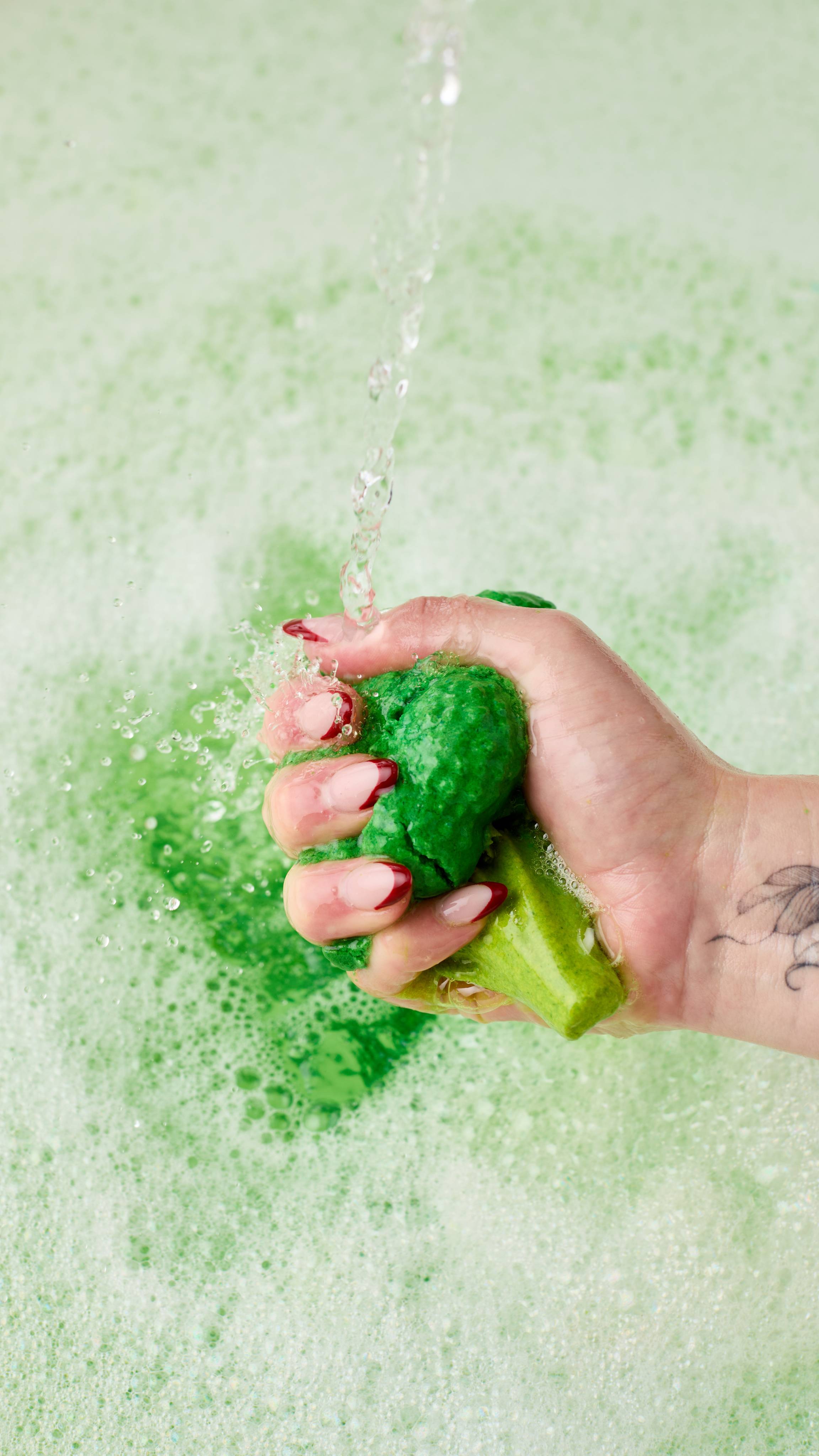 The image shows a hand with long nails, crumbling the vibrant green Sparkly Broccoli bubble bar under a stream of water that splashes off. Delicate green water sits below, covered in a blanket of thick bubbles. 