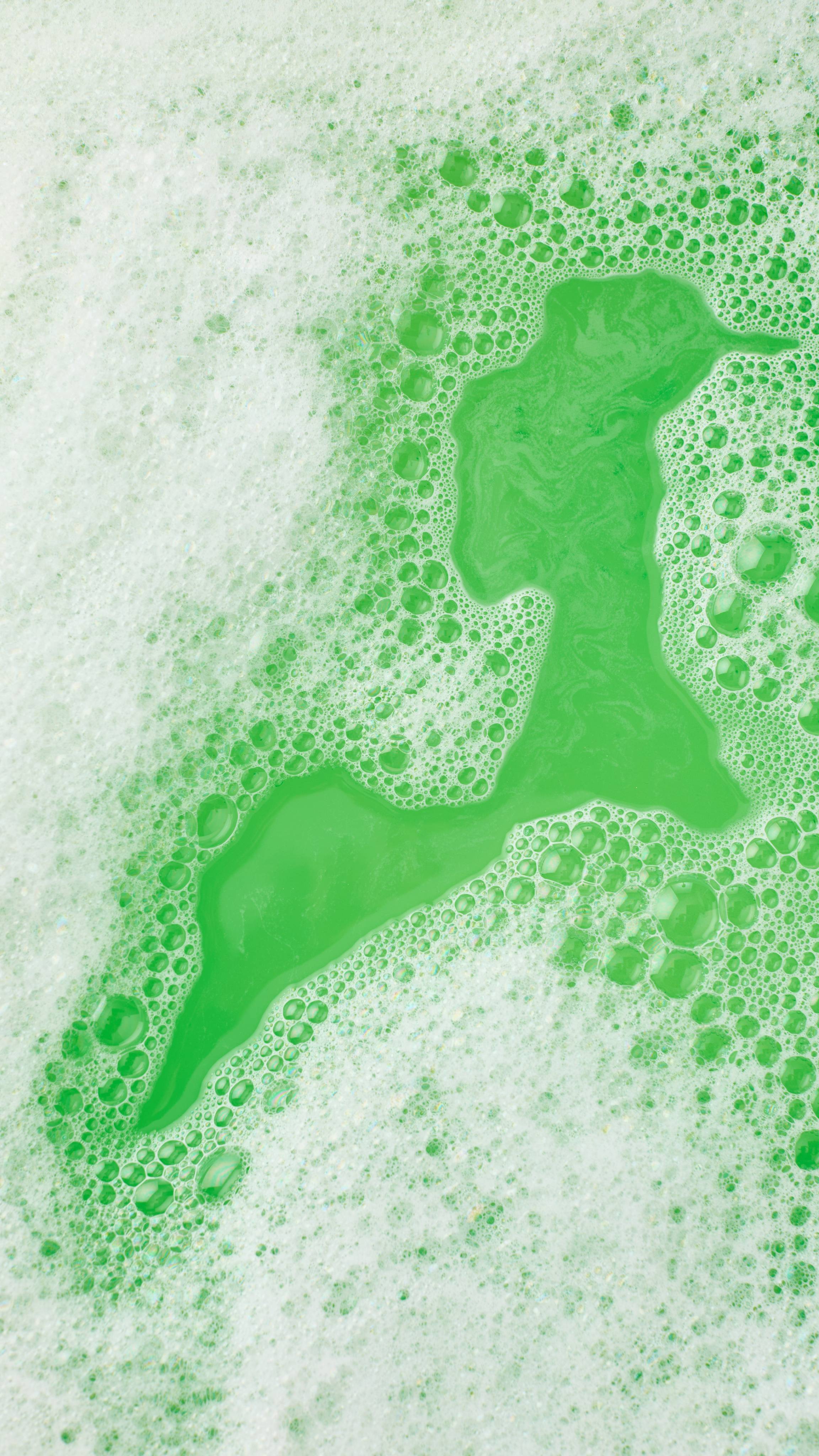 A close-up, aerial image of the bath water after using the Lush, Sparkly Broccoli bubble bar. There is a blanket of thick, fat bubbles with vibrant green bath water visible through a large gap.