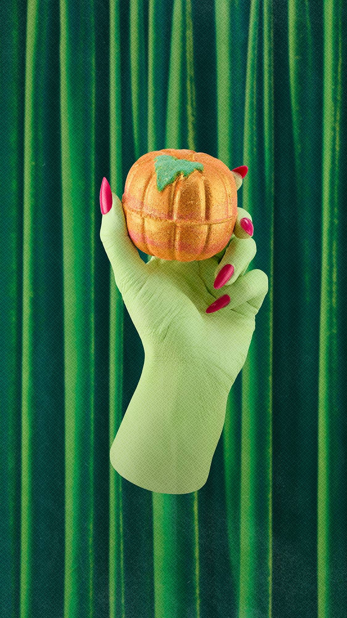 A green hand with long pink nails holds a shimmering, pumpkin-shaped golden-orange bath bomb with a green leaf detail. The hand reaches up against a background of pleated emerald green velvet curtains.