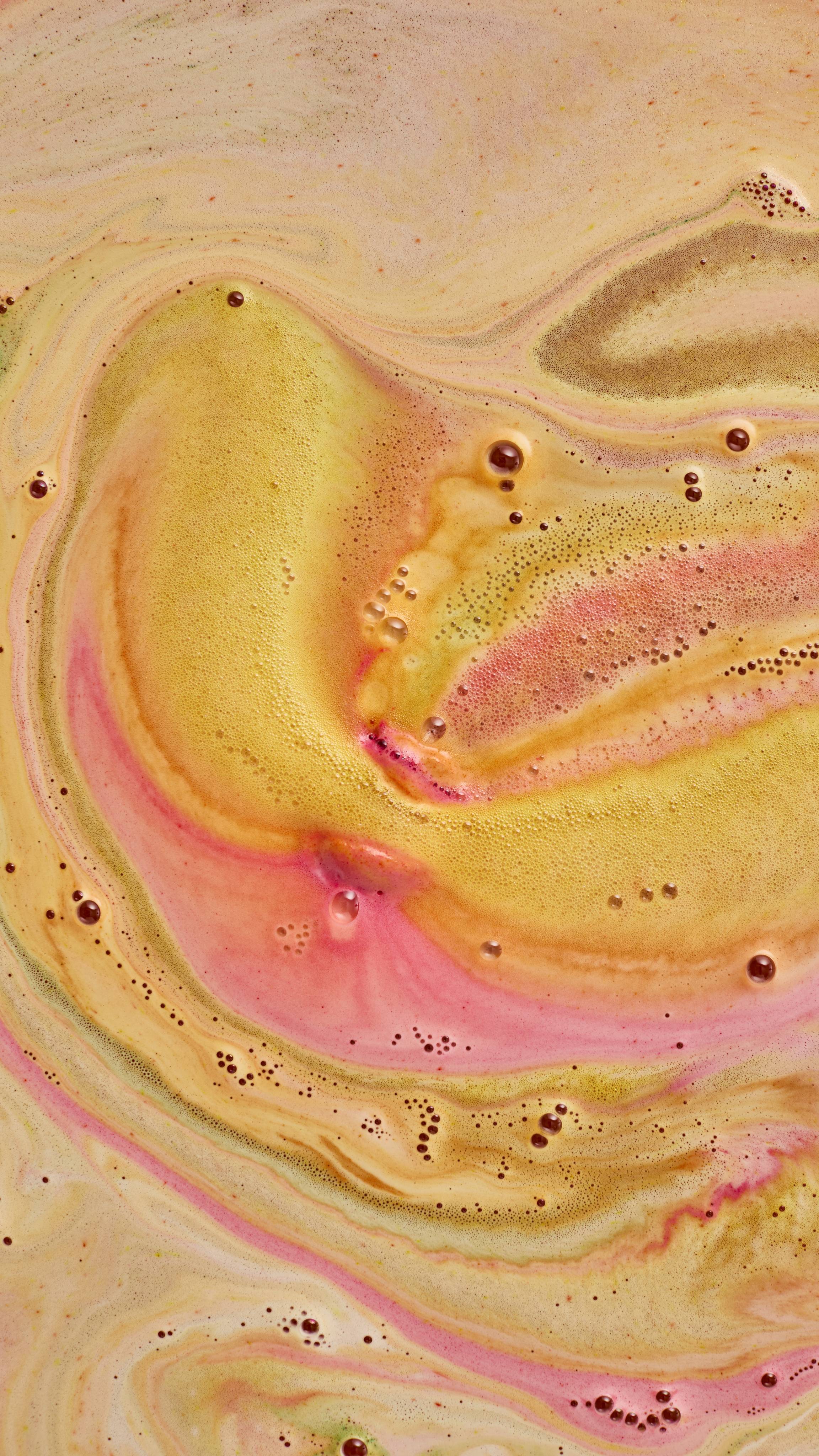 A close-up abstract view of dissolved bath bomb foam, creating swirling patterns of creamy yellow, shimmering gold, soft pink, and peach across the surface of the bathwater with tiny bubbles throughout.
