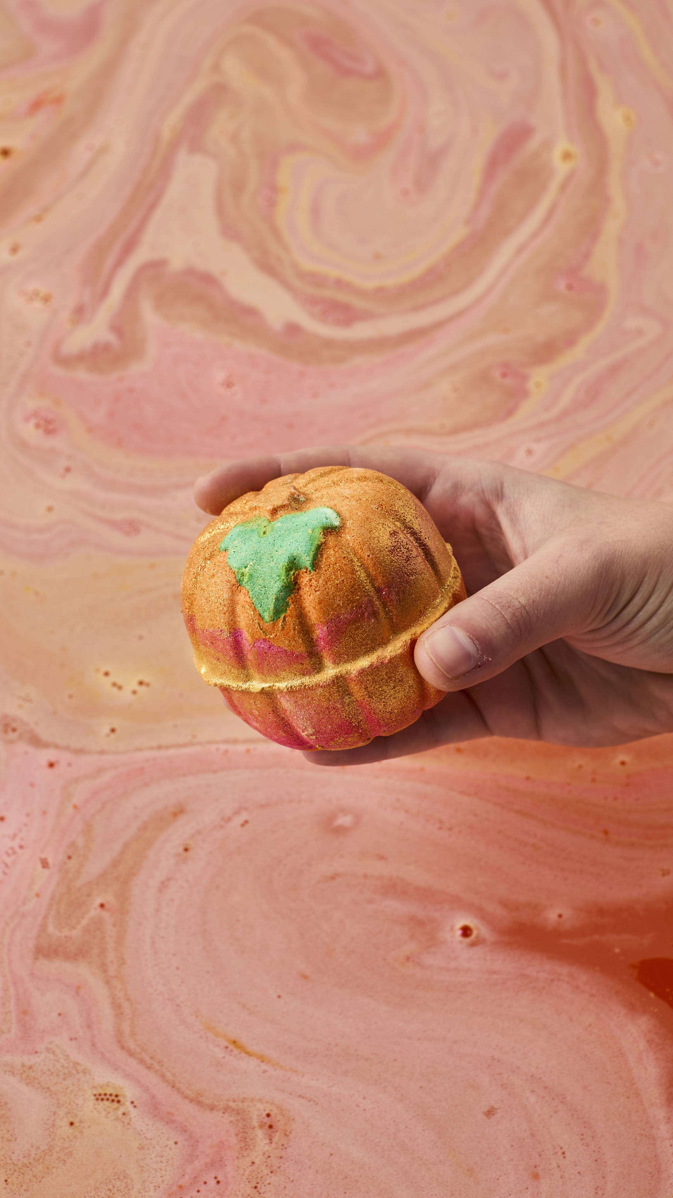 A hand holds a shimmering, pumpkin-shaped golden-orange bath bomb with pink accents and a green leaf detail above bathwater swirling with soft pink and peach foam.