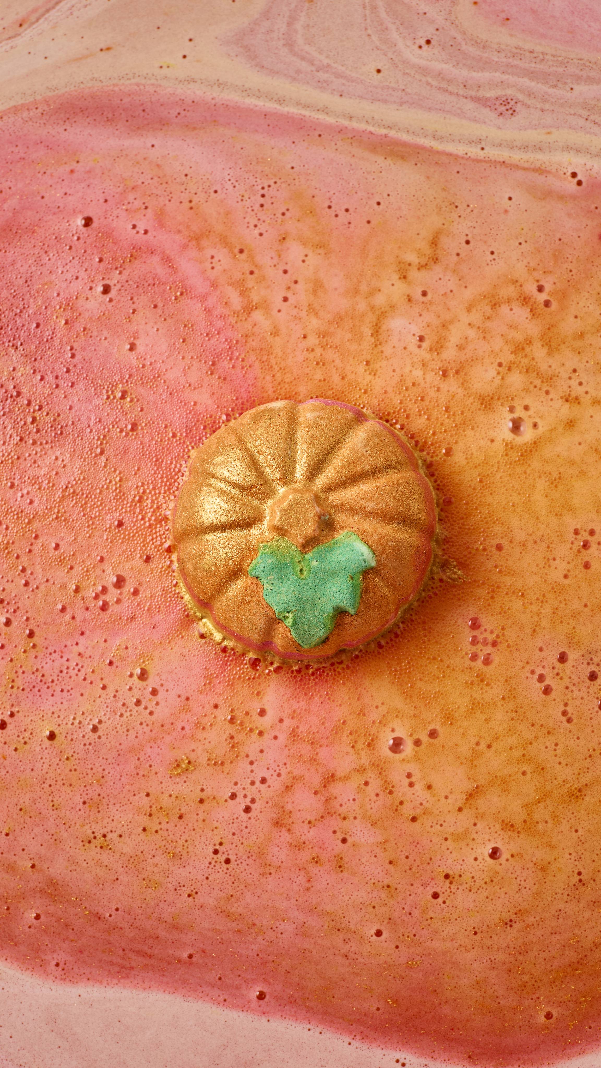 A top-down view of a golden, pumpkin-shaped bath bomb fizzing in the tub, floating amidst swirling streams of sparkling pink, orange, and golden foam on top of the bathwater.