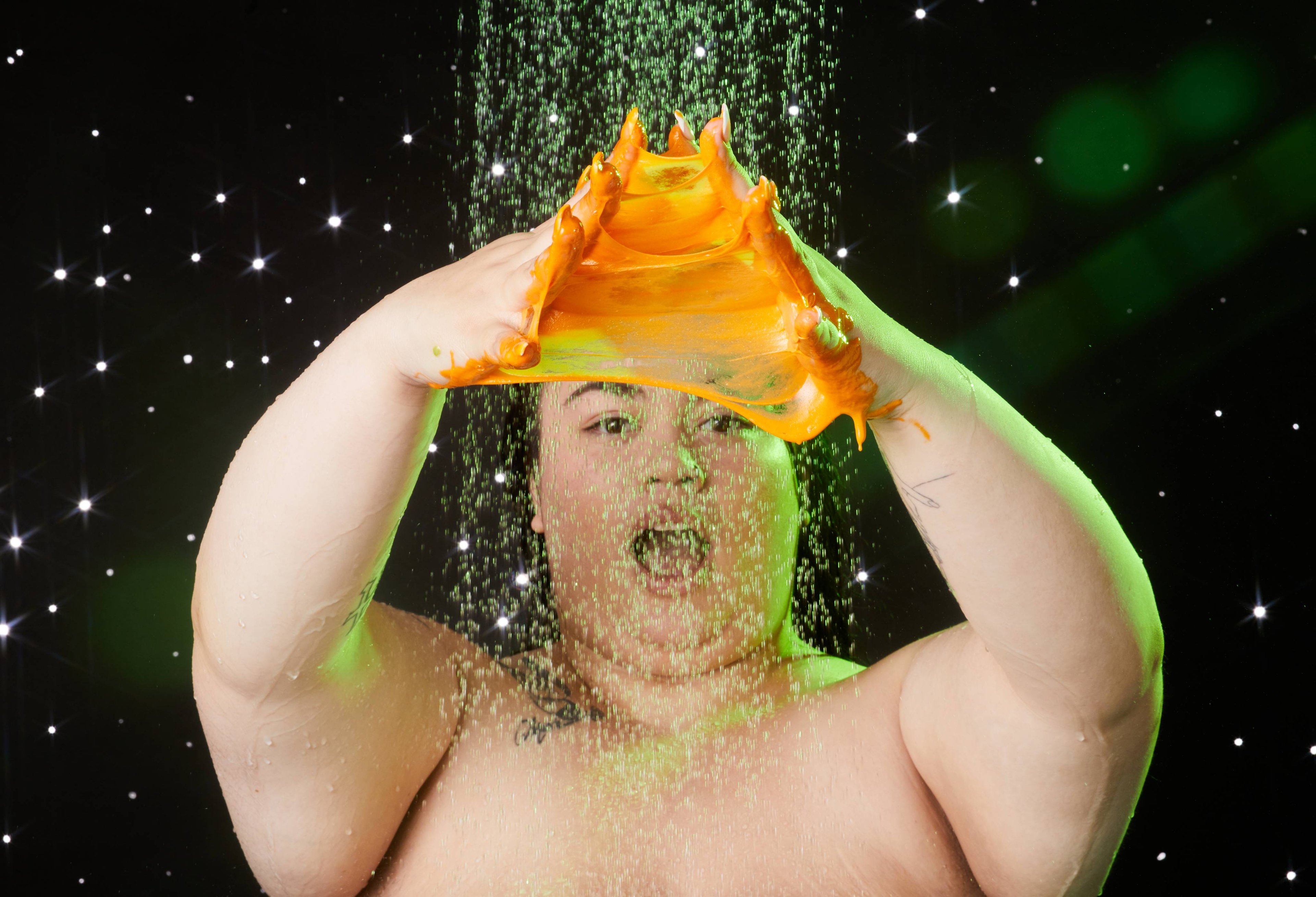 Model stands under the shower with both hands in front of them stretching the product creating a fun, orange, slimy webbing.
