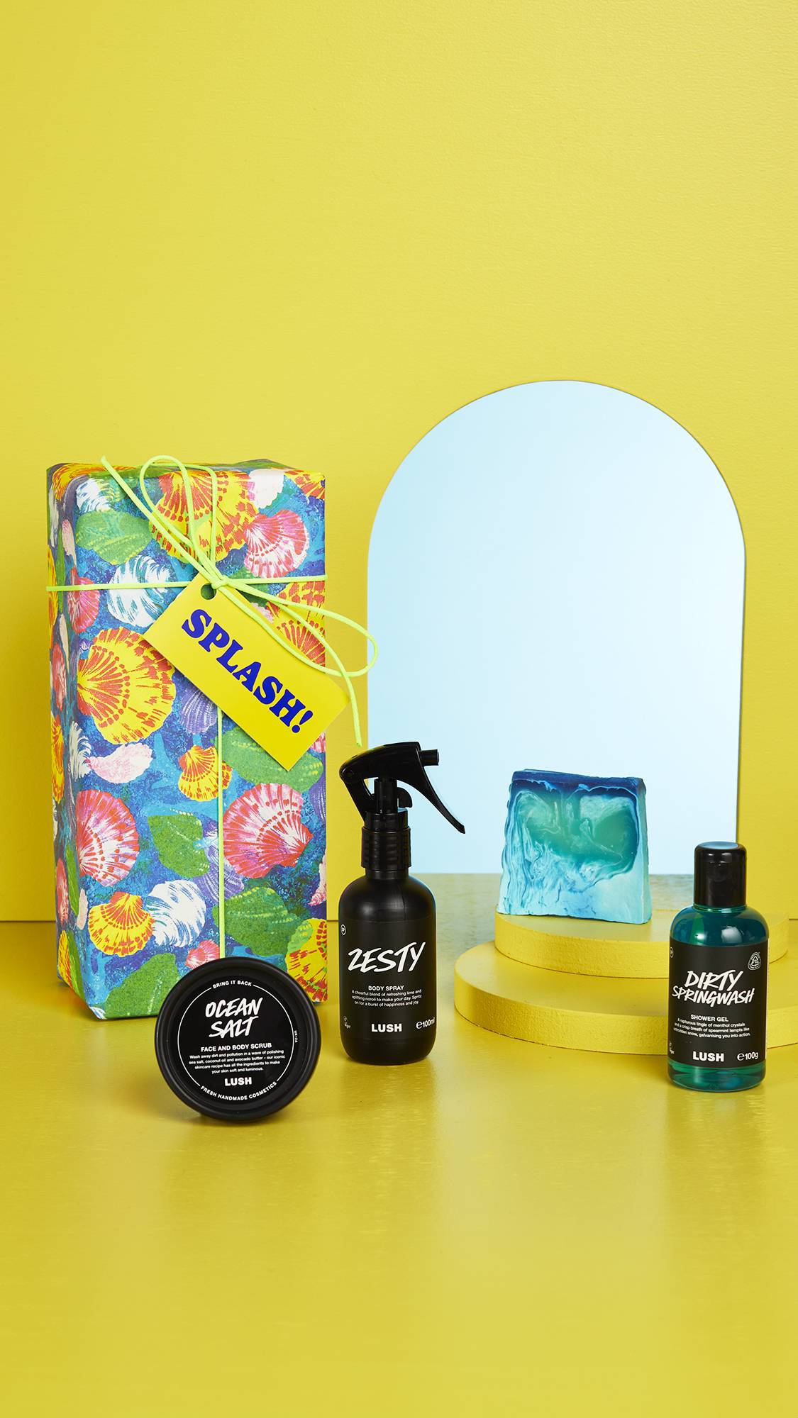 The shell-patterned gift box and the four LUSH products sit on a bright yellow floor and background with an arched mirror in the background.
