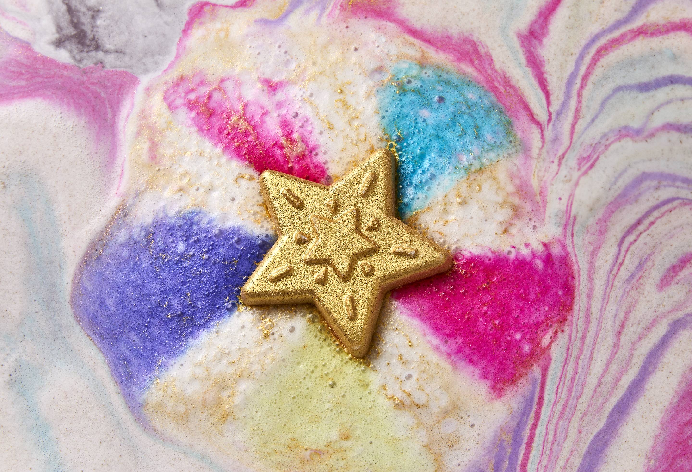 The image shows a golden, five-pointed Star of Wonder bath bomb fizzing in colourful bath water. Vibrant streaks of pink, blue, and purple bleed from the star, creating swirling patterns and shimmering golden foam across the water.