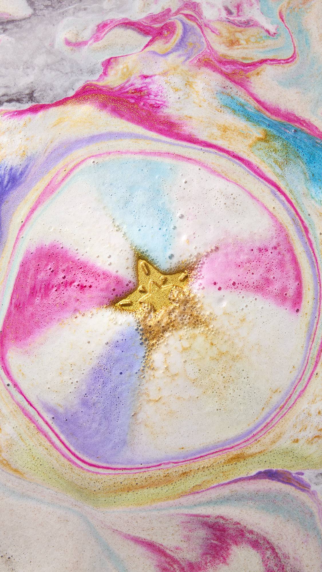 The image shows the golden, five-pointed Star of Wonder product dissolving in colourful bath water. The water forms a vibrant pinwheel of pink, purple, and blue hues, with a core of shimmering golden fizz in the centre.