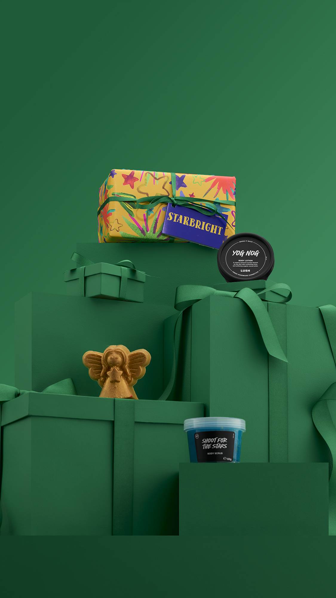 A festive Lush "Starbright" gift set. A large yellow gift box with a red and green star pattern is elevated on a stack of dark green, ribbon-wrapped boxes, with a gold angel-shaped bath product and other items arranged around the set.