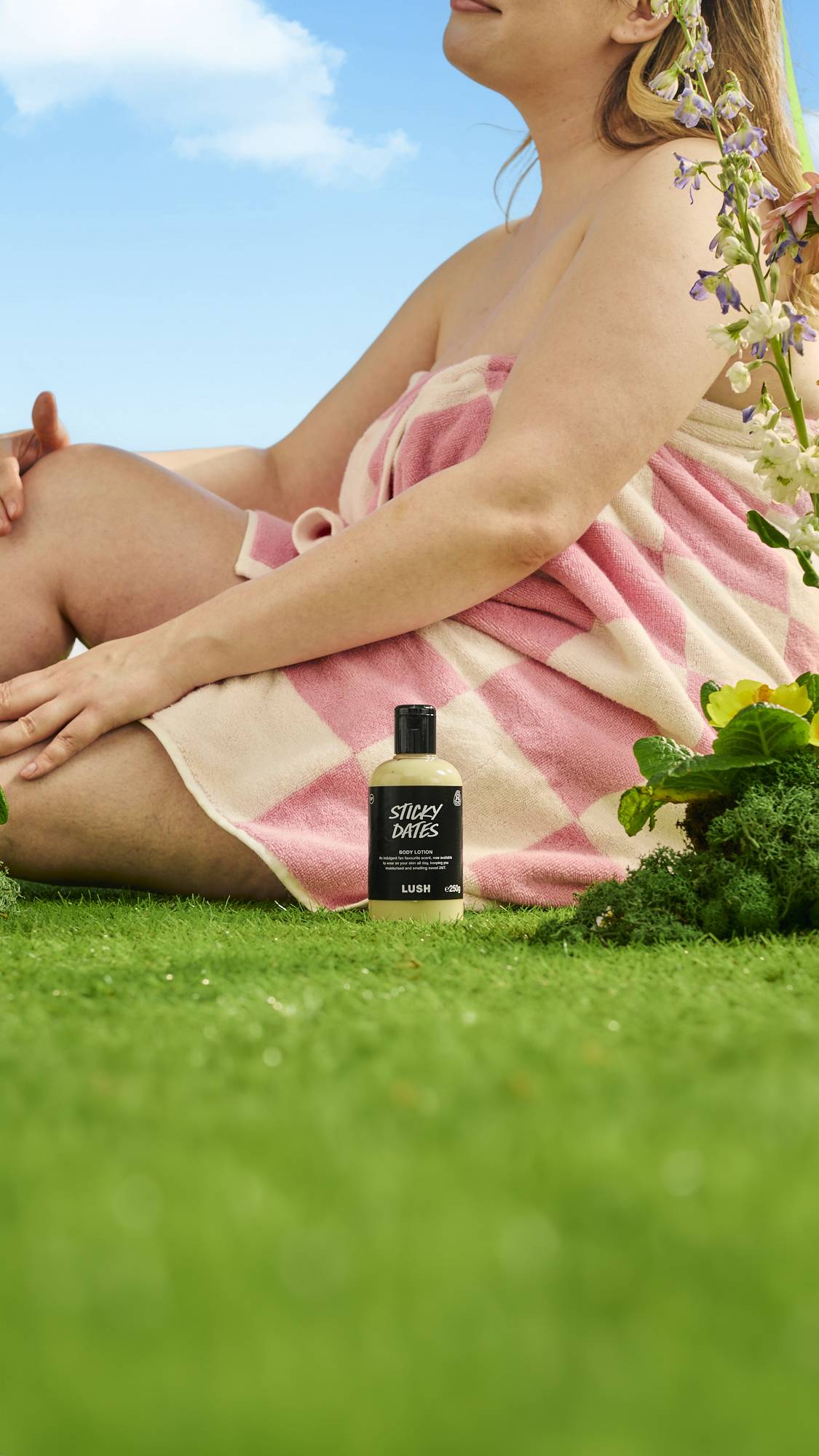 The model is wrapped in a pink and white towel as they sit on the grass under a blue sky while applying the Sticky Dates body lotion to their propped-up knee as the product sits in front. 