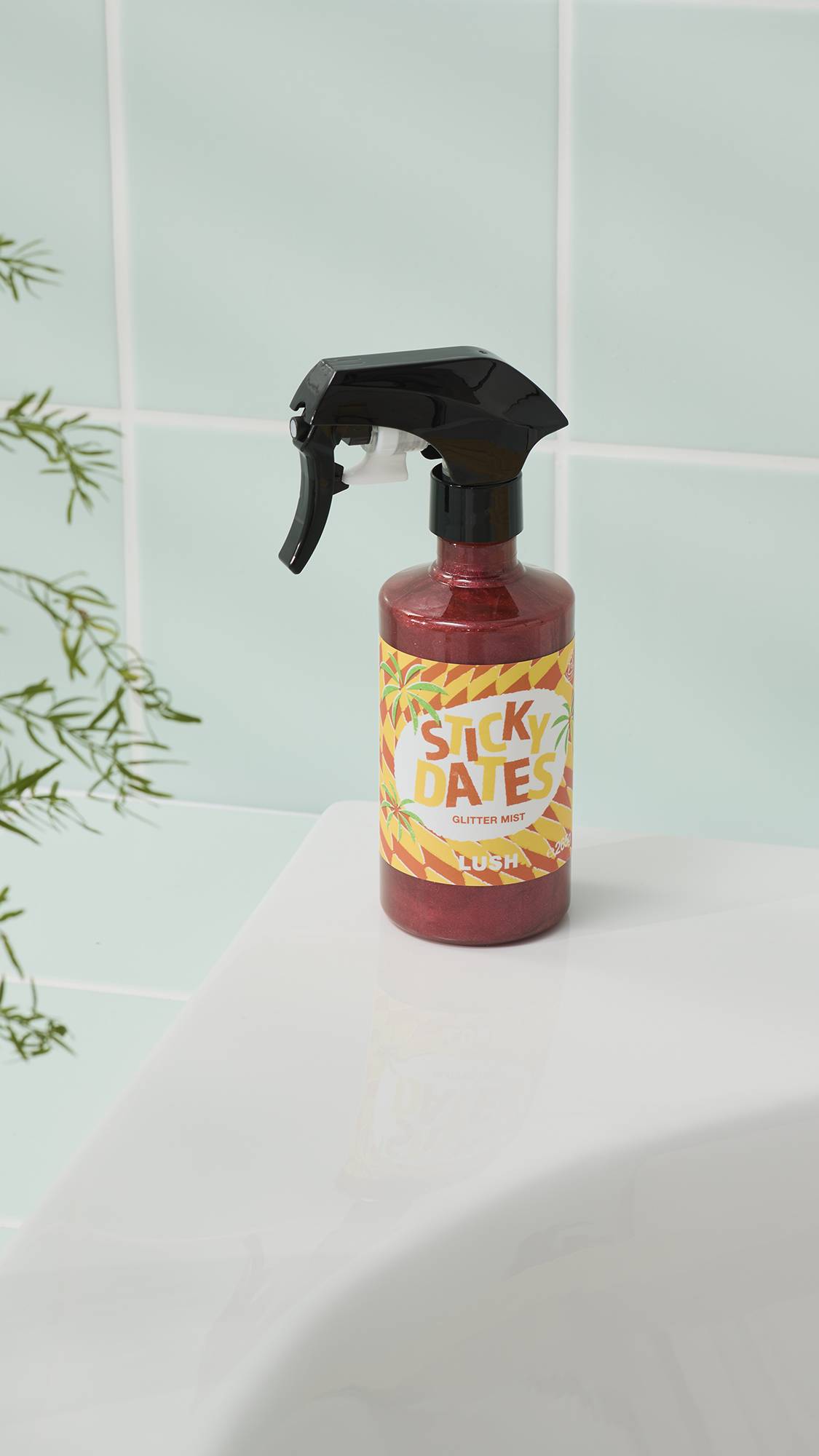 A spray bottle of Lush ‘Sticky Dates’ glitter mist sits on a white surface in front of light blue bathroom tiles. The bottle contains a shimmering, reddish-brown liquid and has a black trigger spray head.