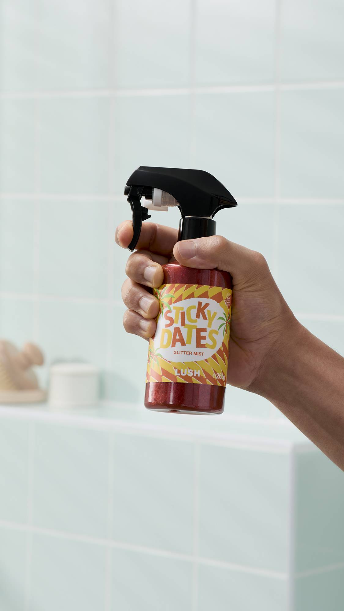 A model's hand holds a bottle of Lush "Sticky Dates" Glitter Mist in a bathroom setting. The bottle contains a shimmering, deep-red liquid and features a black spray nozzle and colourful label. The background shows pale green wall tiles and a shelf.