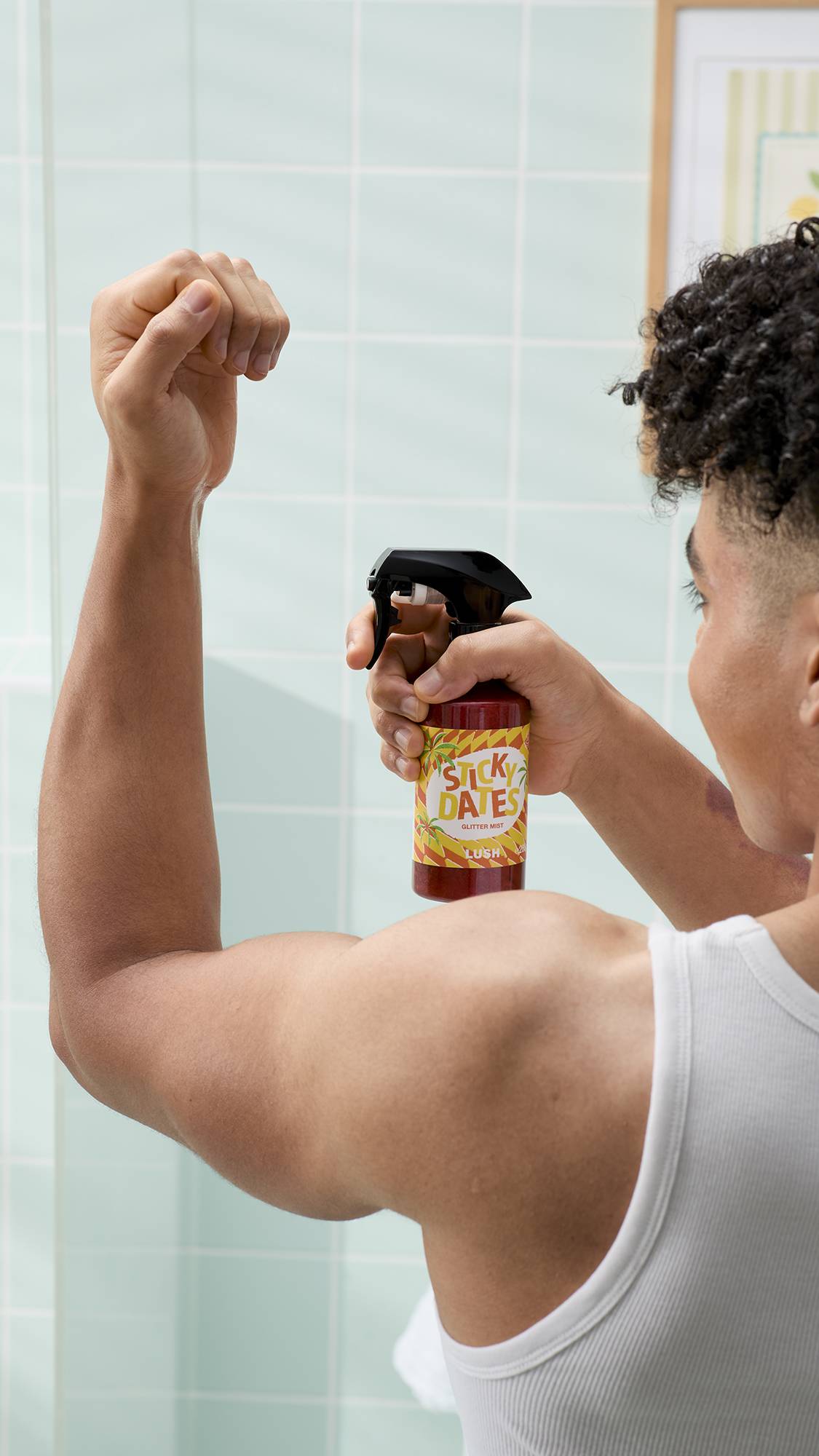 A model with curly dark hair wearing a white vest stands in a bathroom, with their arm lifted. They hold a bottle of Lush ‘Sticky Dates’ glitter mist, pointing the spray nozzle towards their raised arm against a backdrop of light blue tiles.