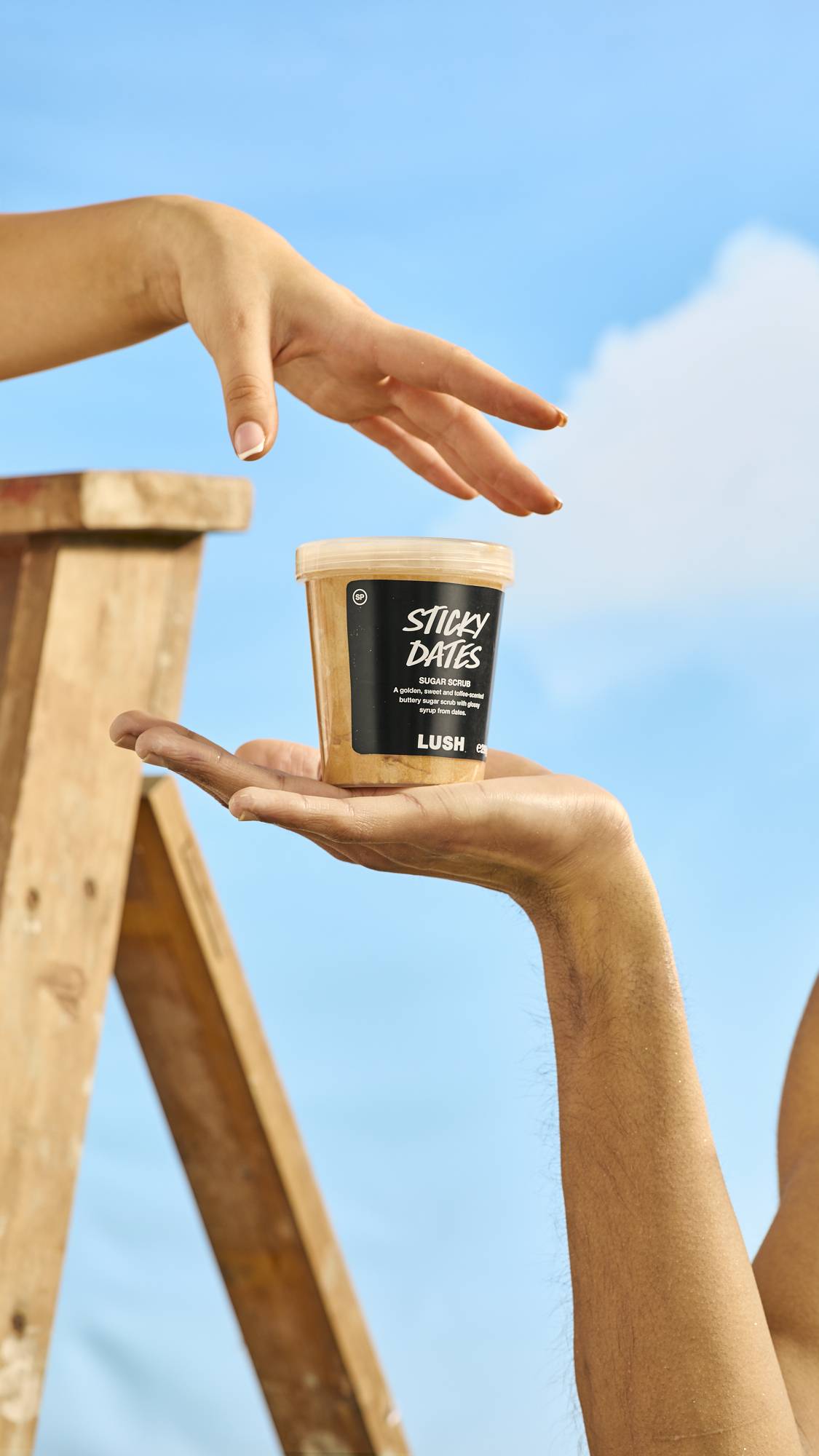 The image shows a close-up of the model's hand out flat while holding the Sticky Dates body scrub lush pot. The sky is blue and another hand is reaching in for the pot. 