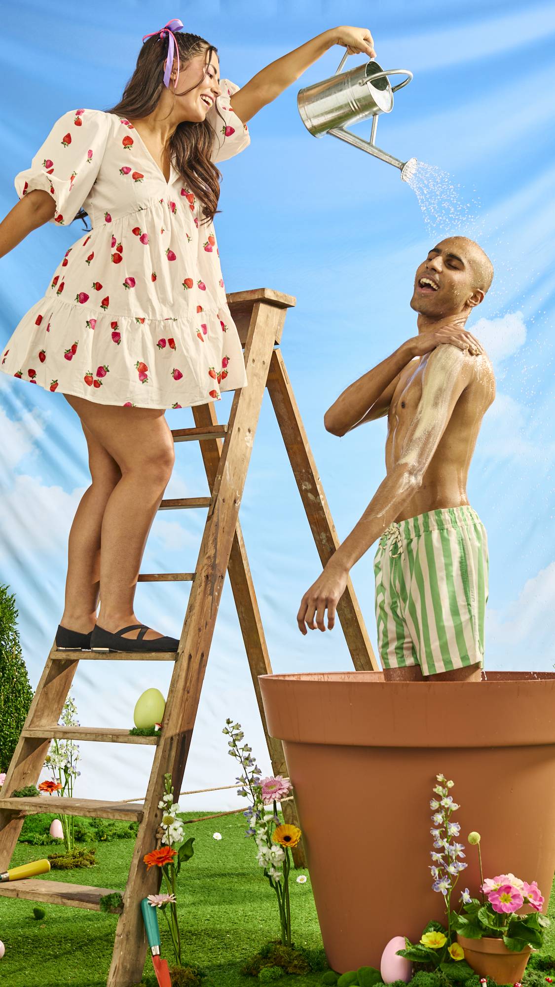 One model is stood on a wooden ladder pouring a watering can on top of the second model who is stood in a giant terracotta pot under the watering can as they use the body scrub. 