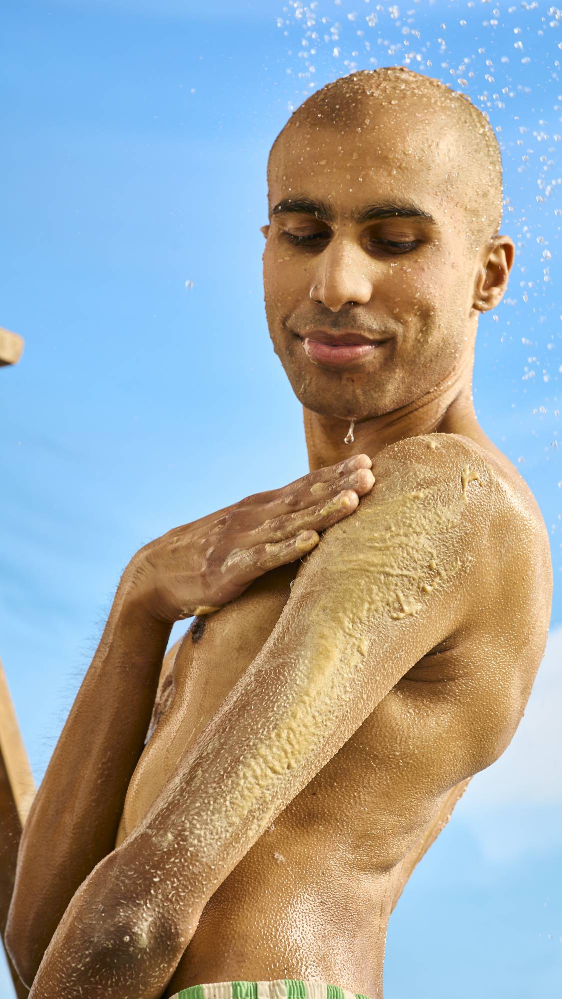 The model is standing under an outdoor shower under a bright blue sky as they exfoliate their upper arm with the Sticky Dates body scrub. 