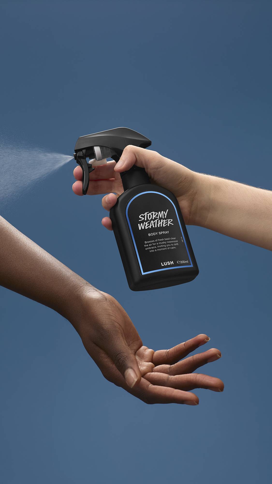 The image shows a light-toned hand spraying a bottle of LUSH 'Stormy Weather' body spray. A warm-toned hand is cupped below, catching the fine mist against a sea-blue background.