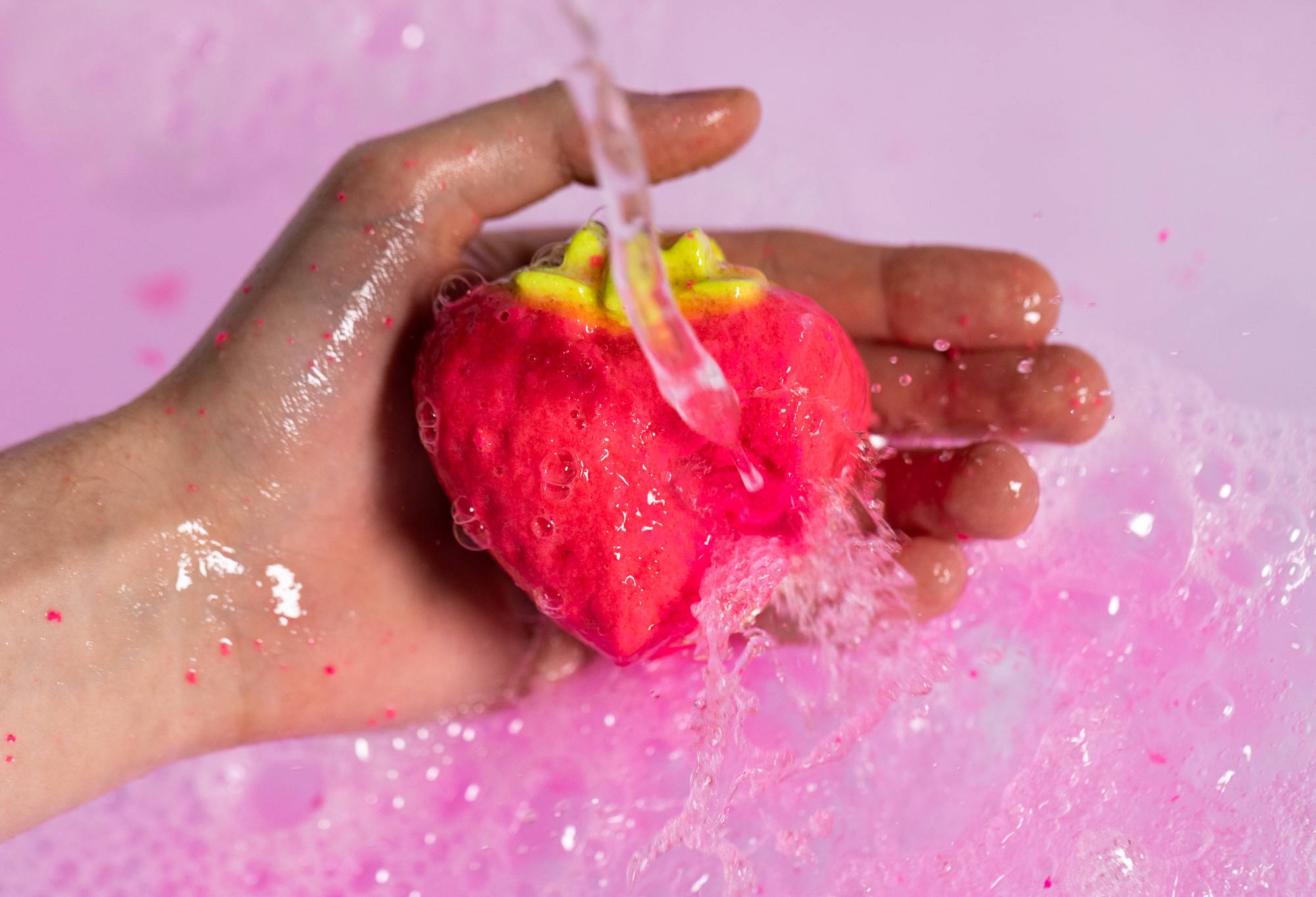 The image shows the model holding a strawberry bubble bar under running water as pink bubbles form below.