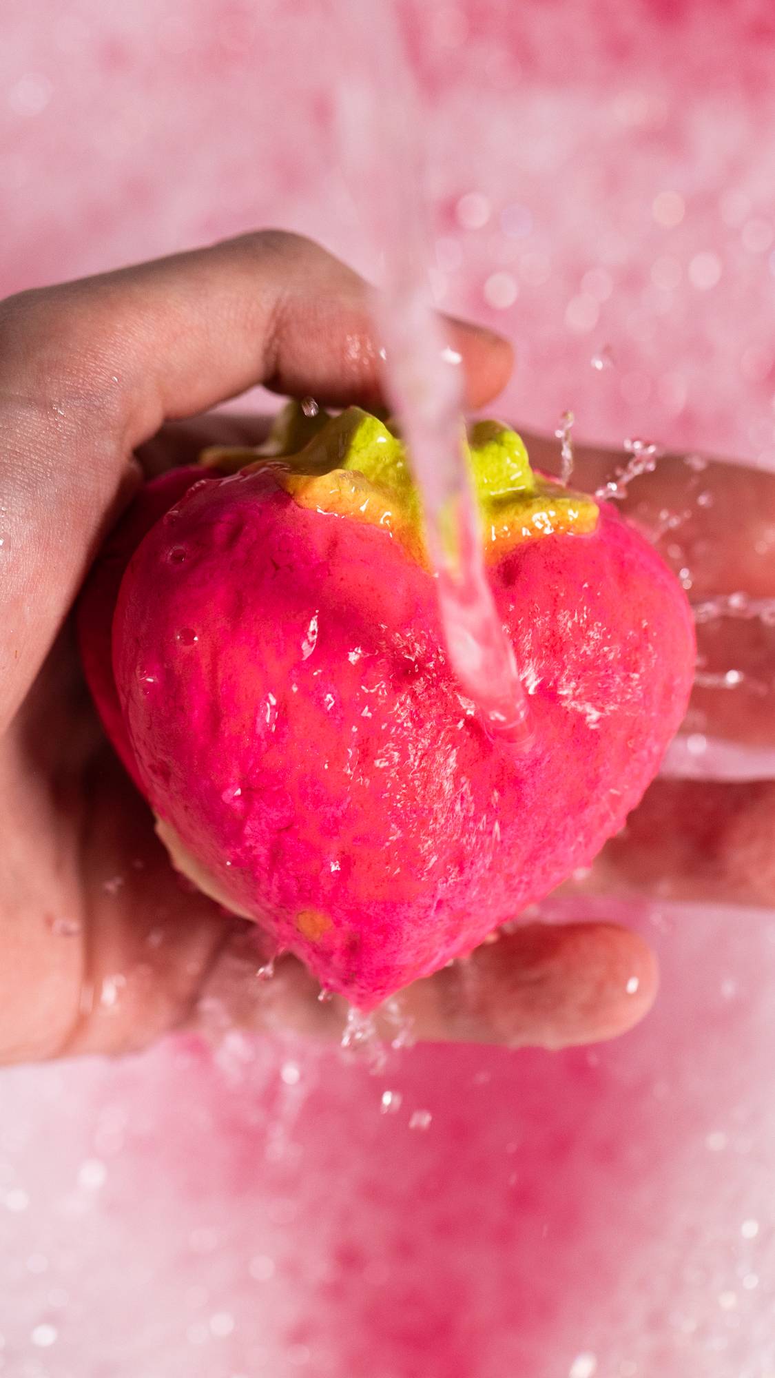 The image shows a close-up of the model holding the Strawberry Crumble bubbleroon under running water creating pink bubbles. 