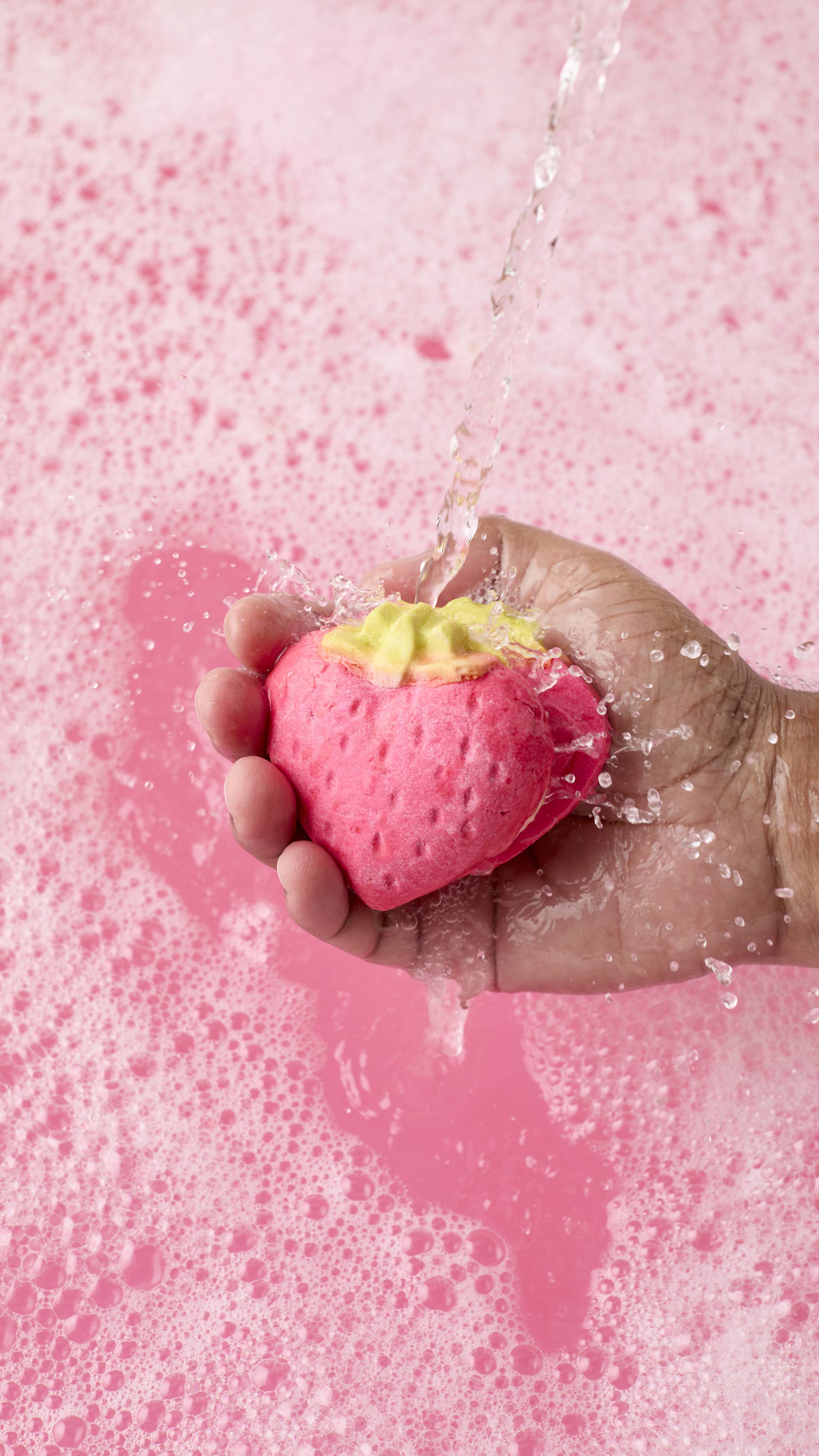 The image shows a hand holding a pink strawberry-shaped bubbleroon bubble bar under a stream of running water. The bubble bar has a green leafy top and is held over a tub of frothy, pale pink bubbles.
