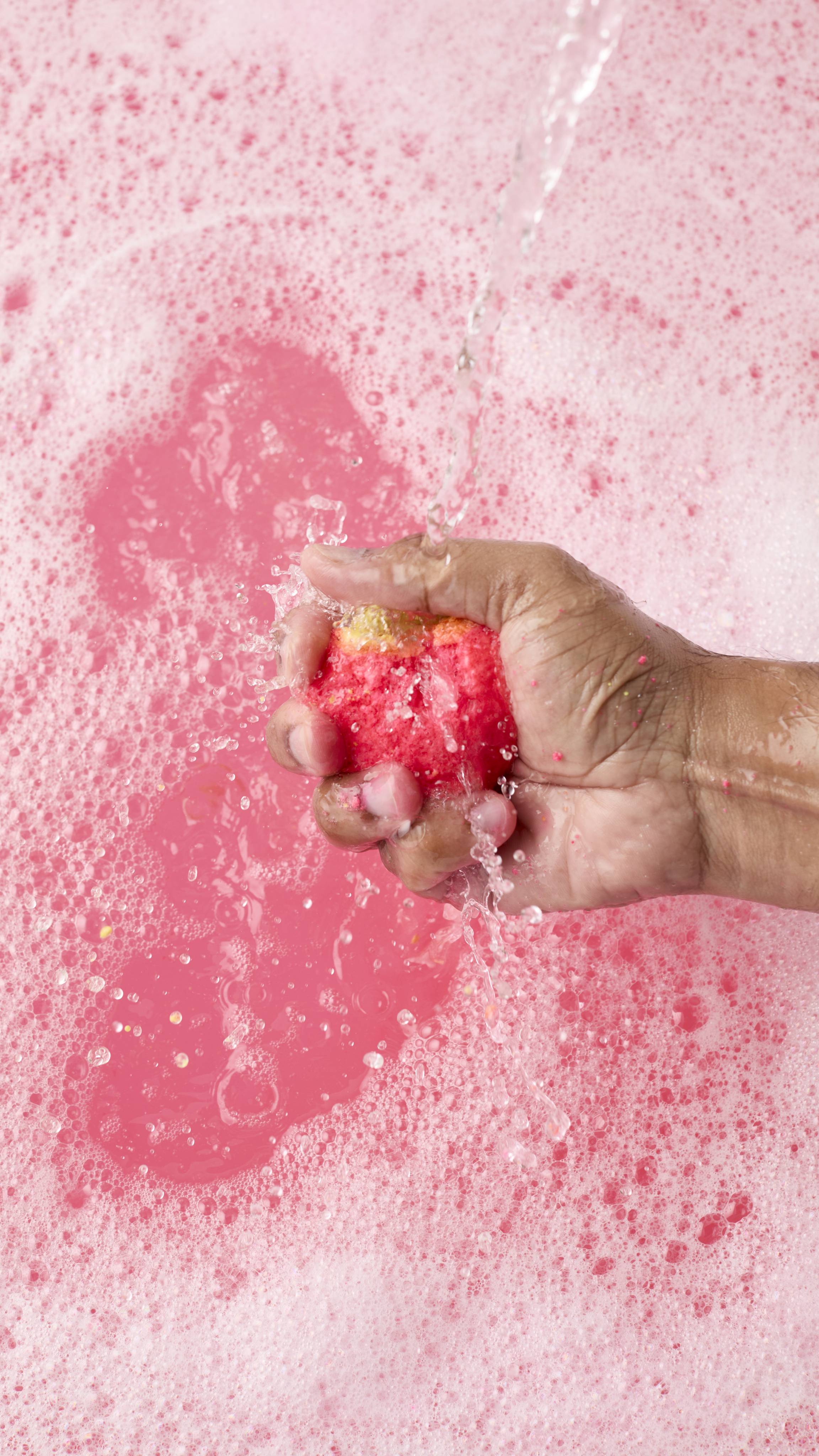 The image shows a hand crumbling a dissolving strawberry-shaped bubbleroon bubble bar under a stream of water. The bubble bar releases bright pink pigment and foam into a tub filled with pale pink bubbles.