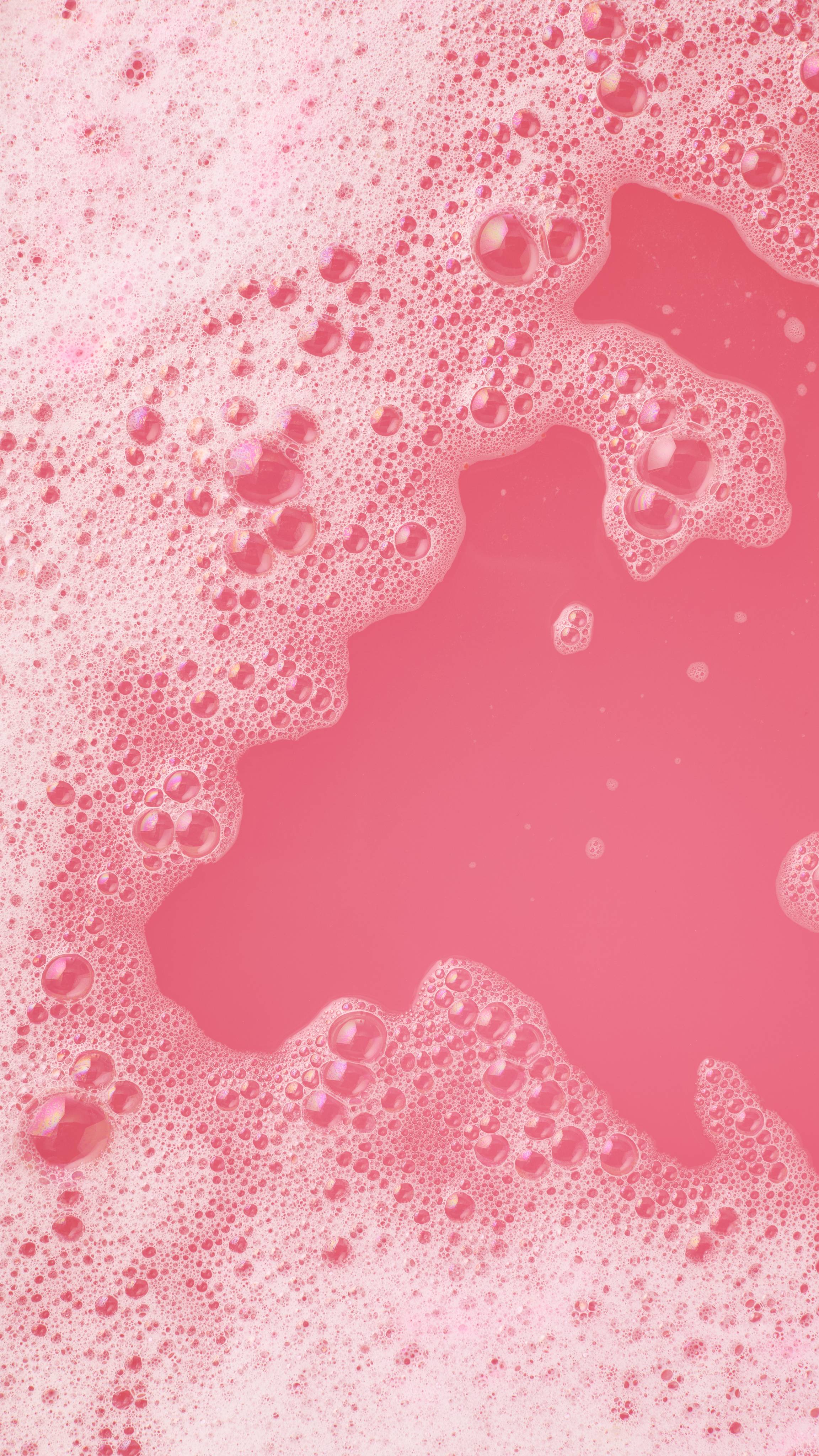 An aerial view of thick, pale pink foam and bubbles floating on top of deep pink bathwater. The bubbles form an irregular, frothy border around a clear patch of pink water, created by the Strawberry Crumble Bubbleroon.