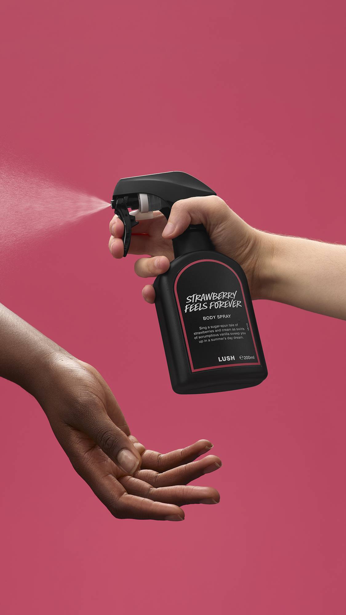The image shows a light-toned hand spraying a bottle of LUSH 'Strawberry Feels Forever' body spray. A warm-toned hand is cupped below, catching the fine mist against a deep, rose-pink background.