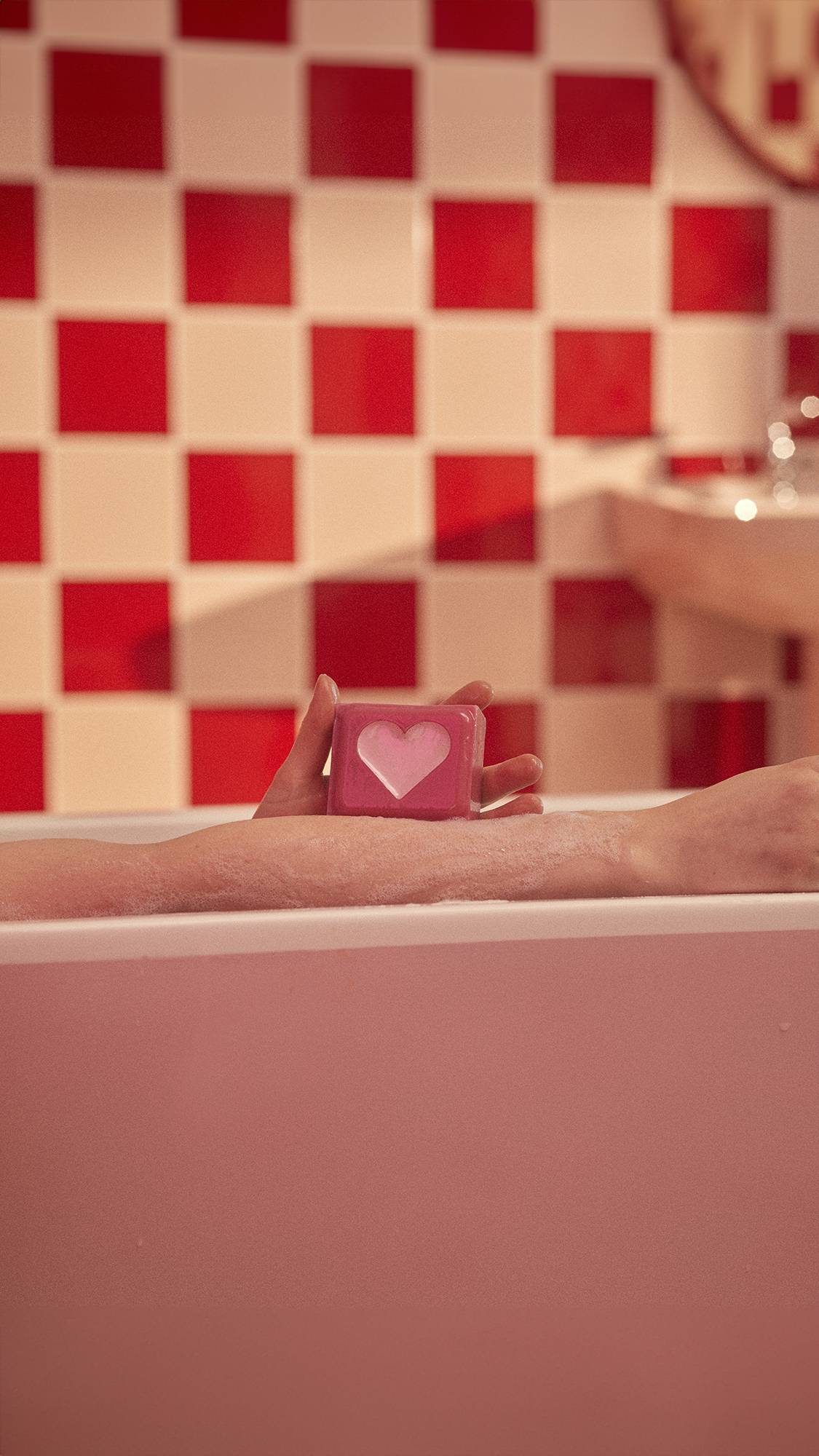 Image shows a tiled bathroom with a pink bath. A forearm lays across the side of the bath as the Strawberry Heart soap is held above.