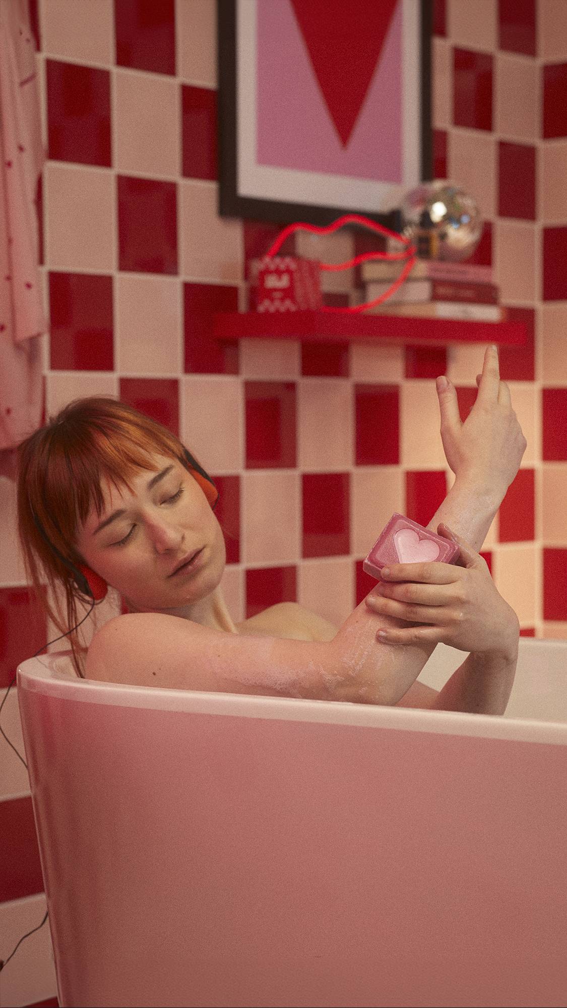 Model is lying in the bathtub in a red and pale pink tiled bathroom as they lather their forearm with the Strawberry Heart soap block. 