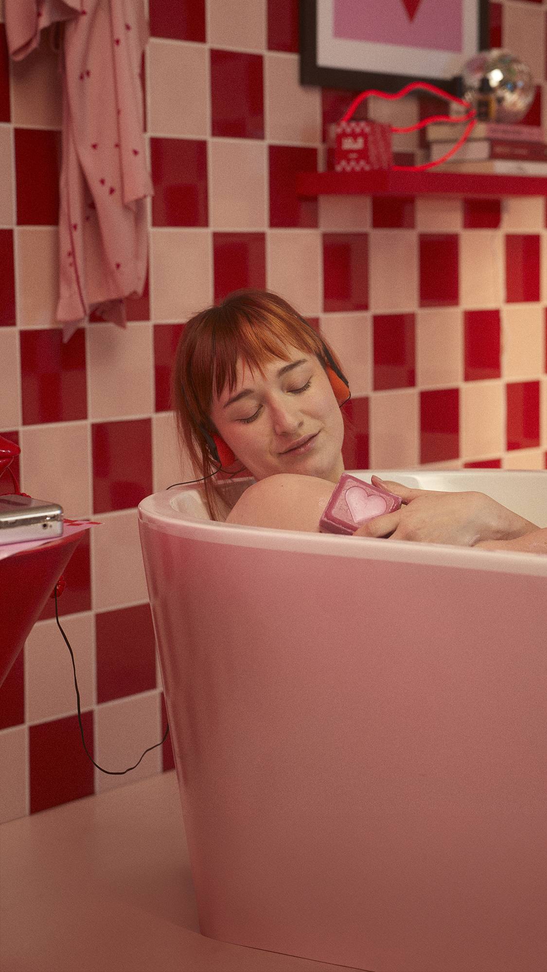 Model is lying in the bathtub in a red and pale pink tiled bathroom as they lather their upper arm with the Strawberry Heart soap block. 