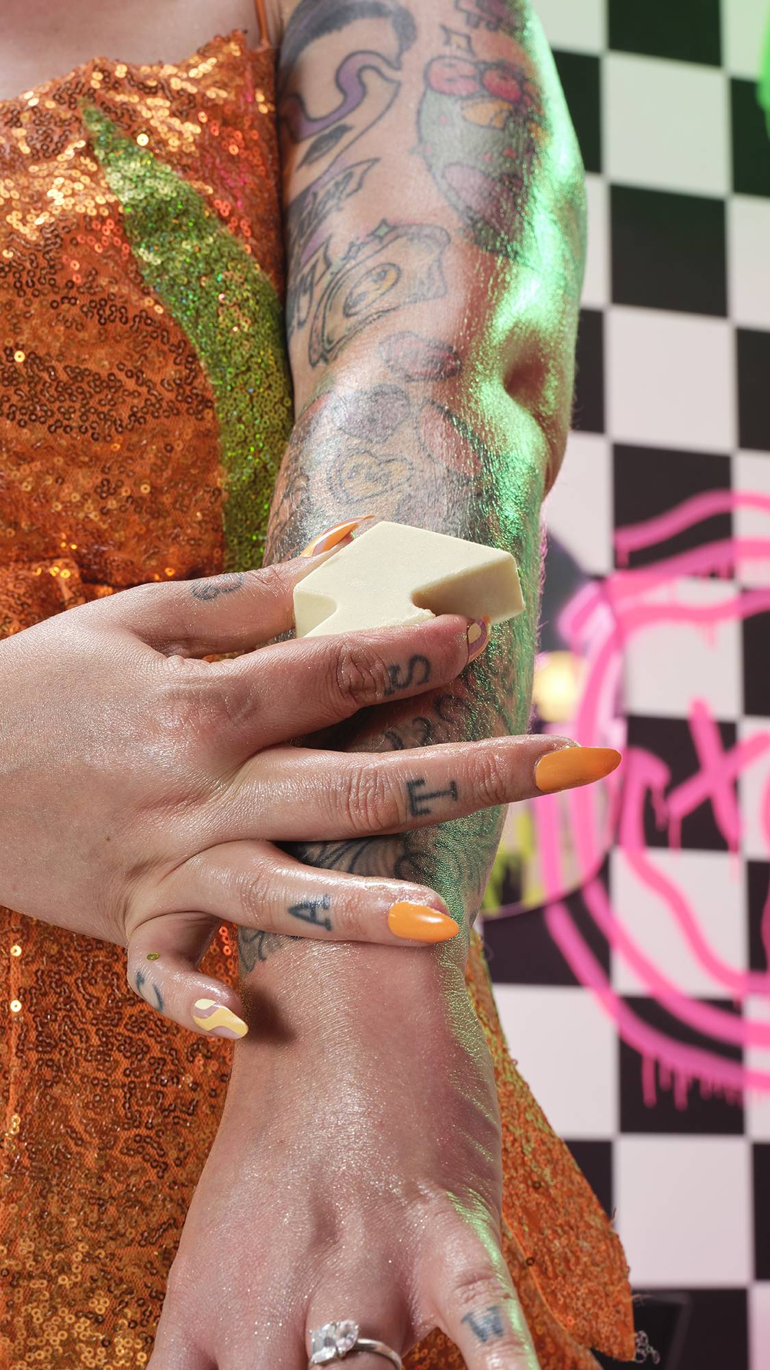 A close-up of the model smoothing the massage bar over their arm. They have an orange sequin dress on a checkered background.