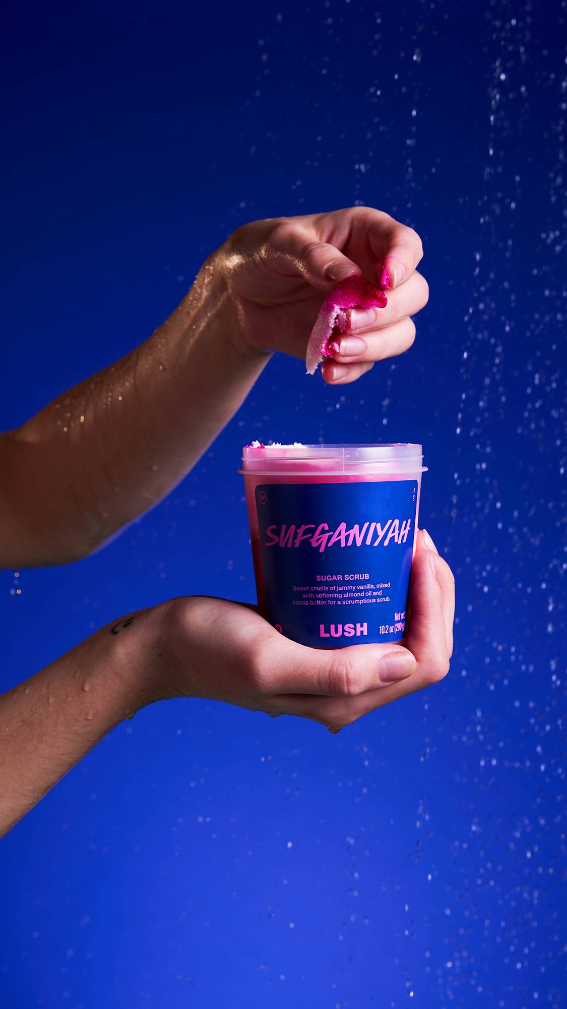 Two hands hold a transparent tub of Sufganiyah Sugar scrub against a deep blue background. The product's interior shows the vibrant white and magenta swirl. The top hand is taking a pinch out under a spray of water.