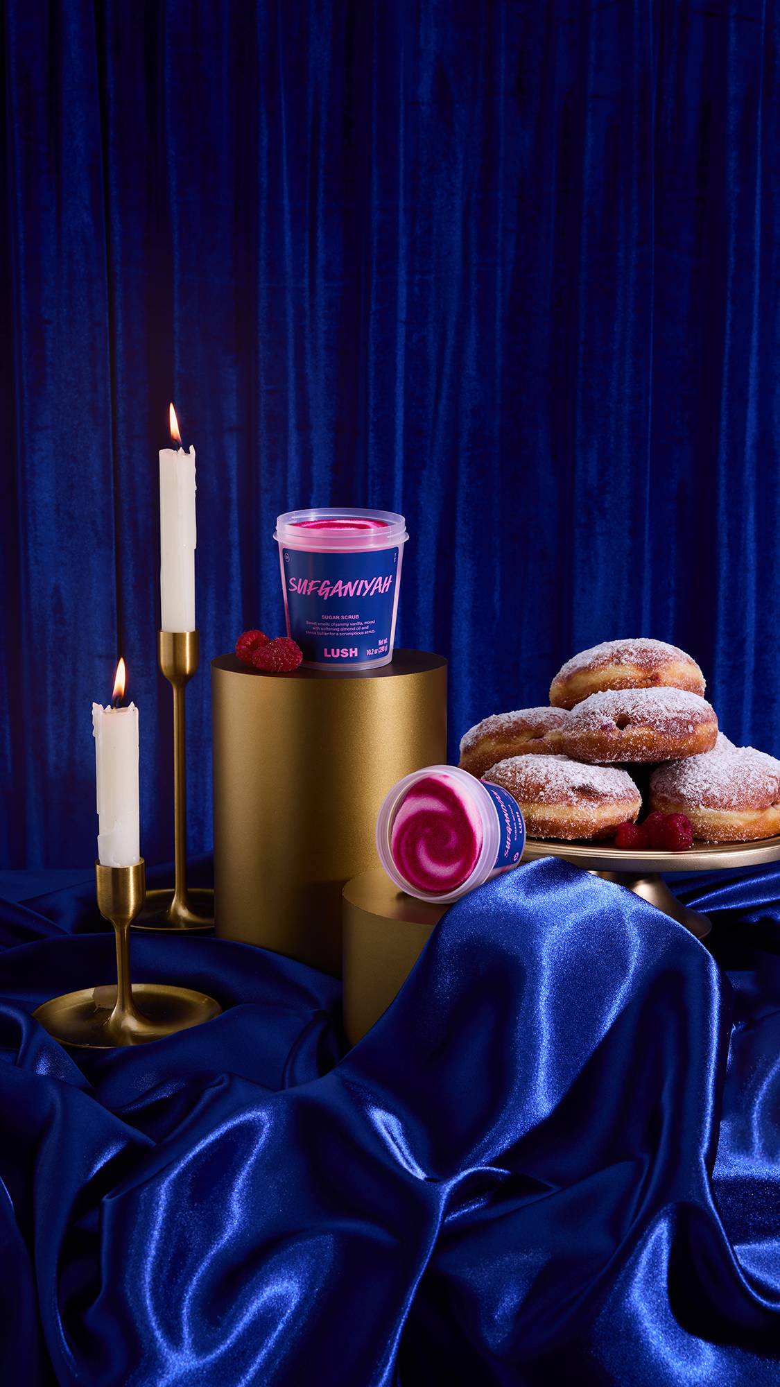 A still life featuring the Sufganiyah Sugar scrub against royal blue velvet and satin. The open scrub shows a white and magenta swirl, displayed with powdered donuts, lit candles in gold holders, and gold cylindrical props.