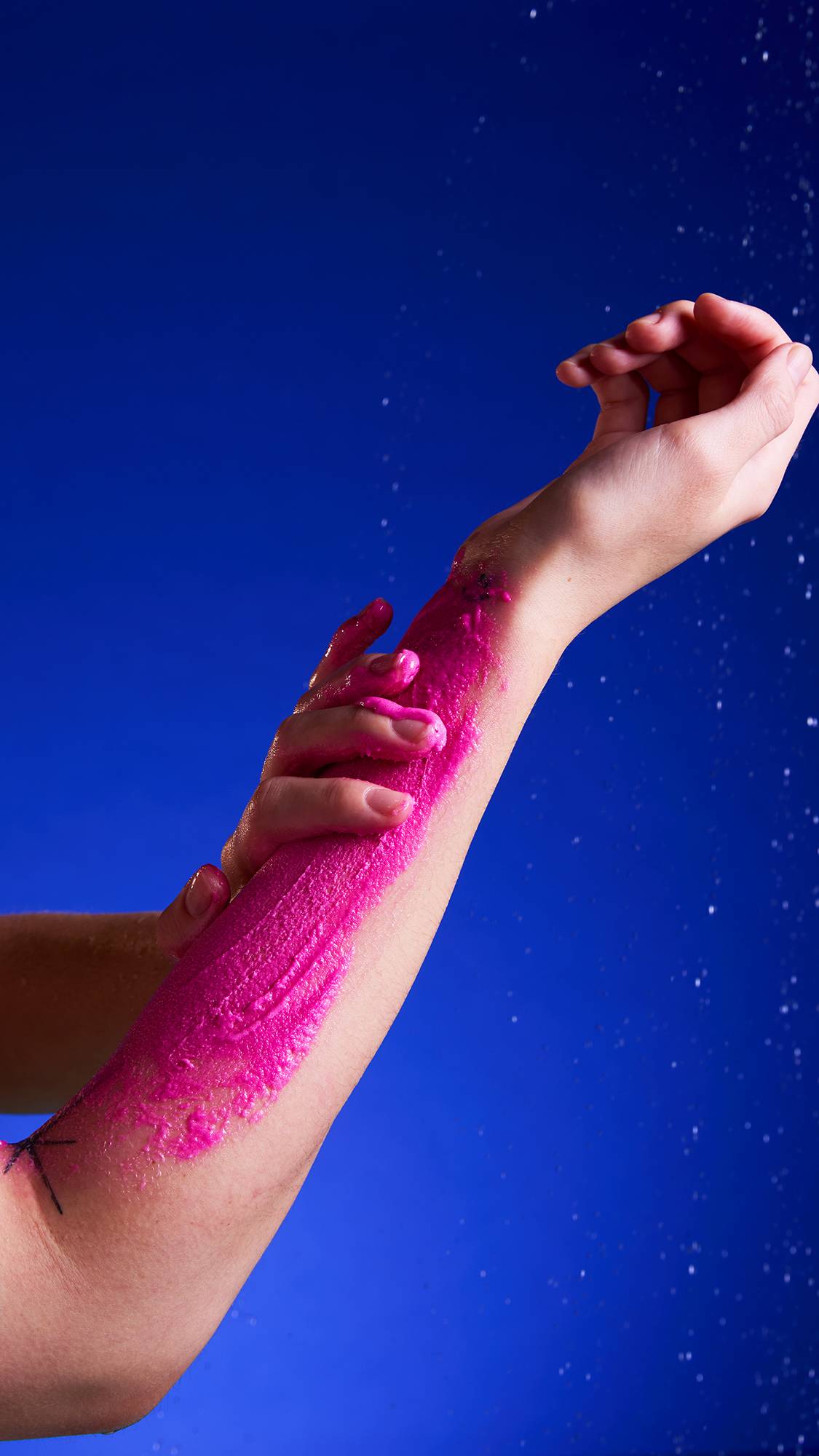 Two hands rub a thick, bright magenta layer of Sufganiyah Sugar scrub onto a forearm. The scrub appears wet, and the scene is under a light spray of water, all set against a deep blue background.
