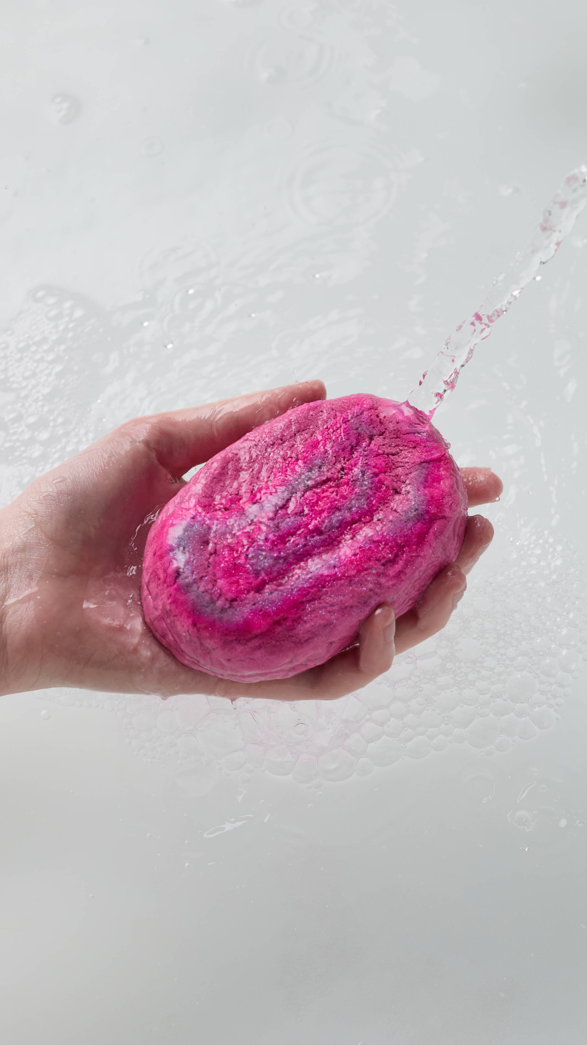 A close-up of the model's hand reaching in from the left while holding the vivid, pink, Lush Sugar Plum Sparkle bubble bar under running water. Delicate white bubbles begin to form on the surface of the bath water below. 