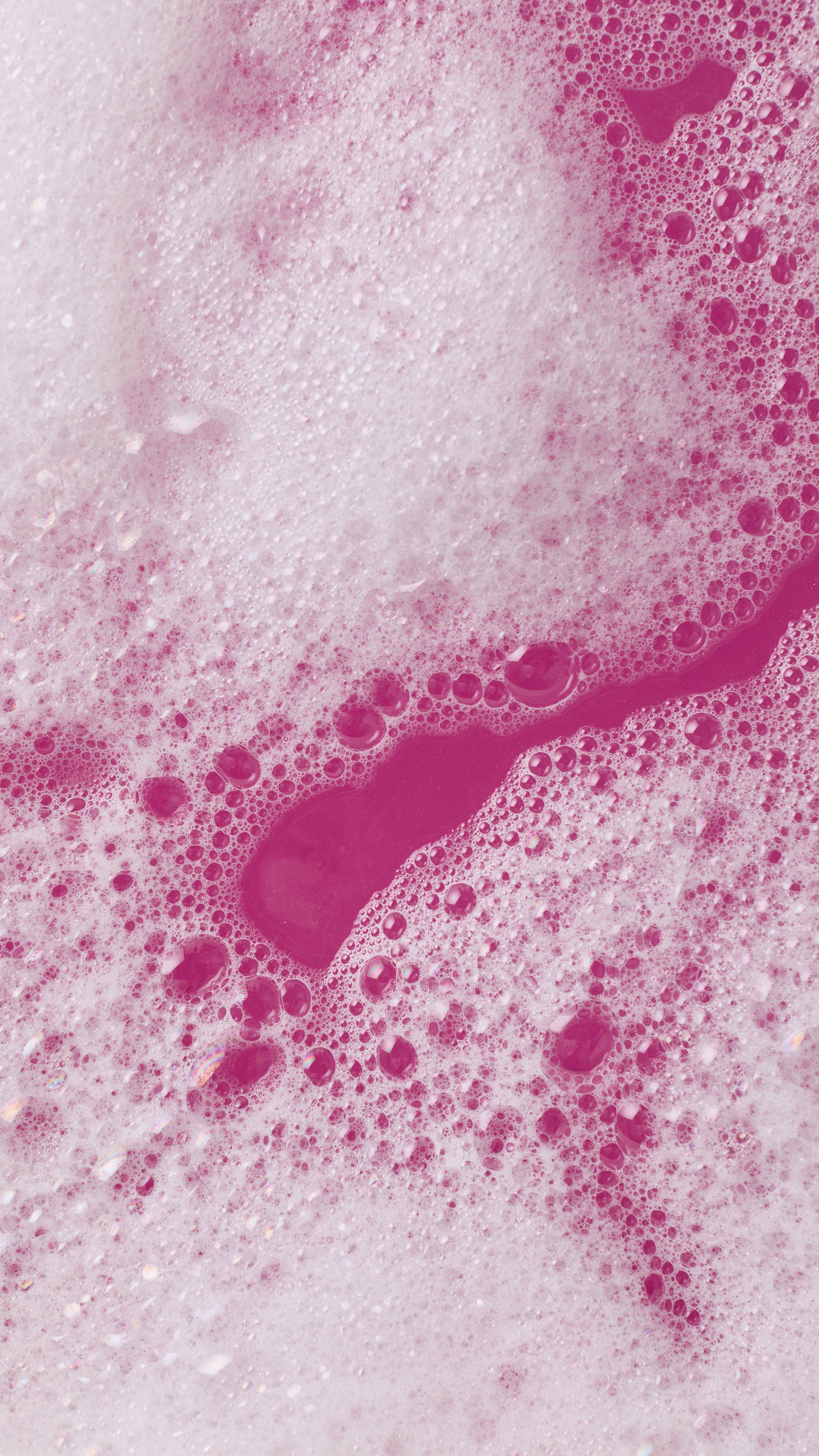 A close-up, aerial image of the bath water after using the Lush, Sugar Plum Sparkle bubble bar. There is a blanket of thick, fat bubbles with bright, bubblegum-pink bath water visible through a large gap.
