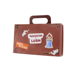 Suitcase. A brown cardboard gift box shaped like a suitcase with a carry handle. The front features 'PADDINGTON x LUSH' branding, an illustration of Paddington Bear in a towel, and a 'WANTED ON VOYAGE' sticker.