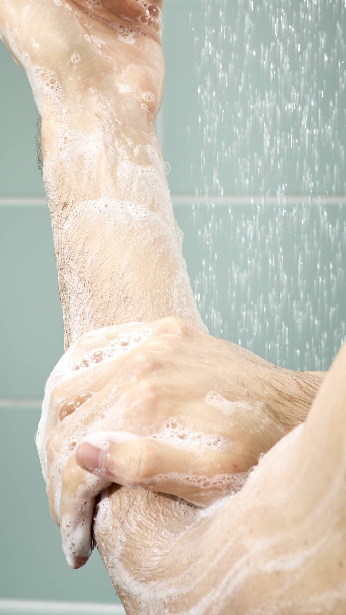 A close-up of a model under a shower, using their hands to massage white, soapy lather along their forearm. Bubbles and water droplets are visible on the skin against a backdrop of light blue tiles and falling water.