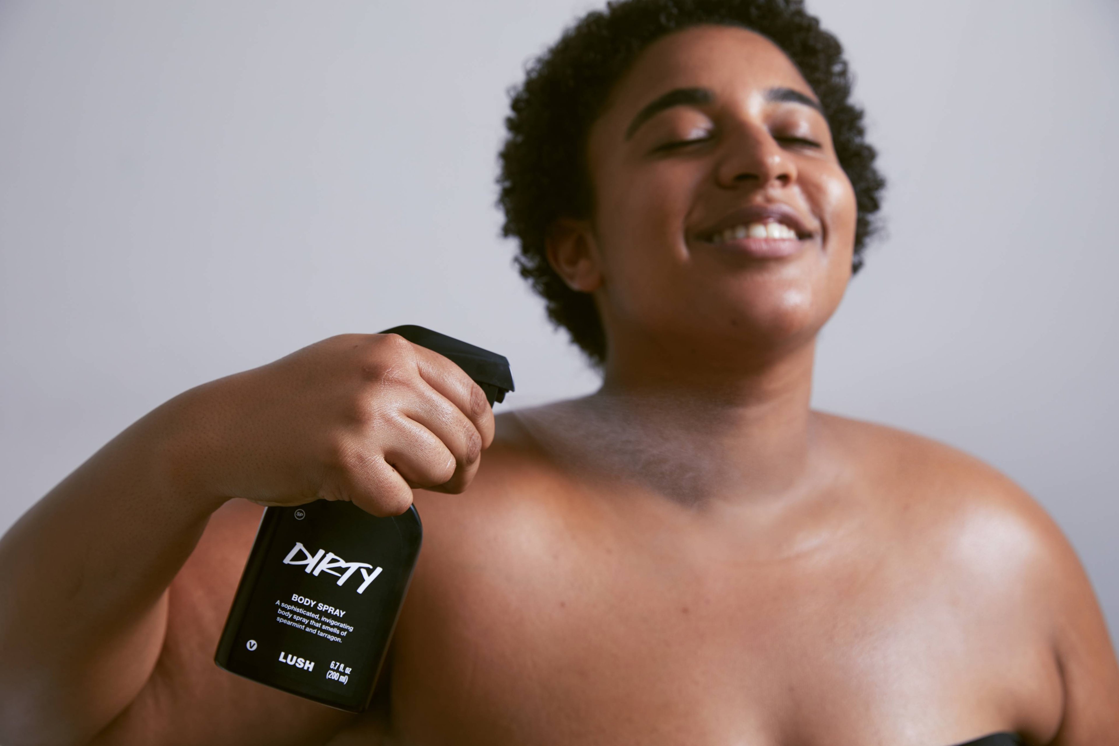 Dirty body spray is sprayed up into the air, in front of shirtless model who's smiling.