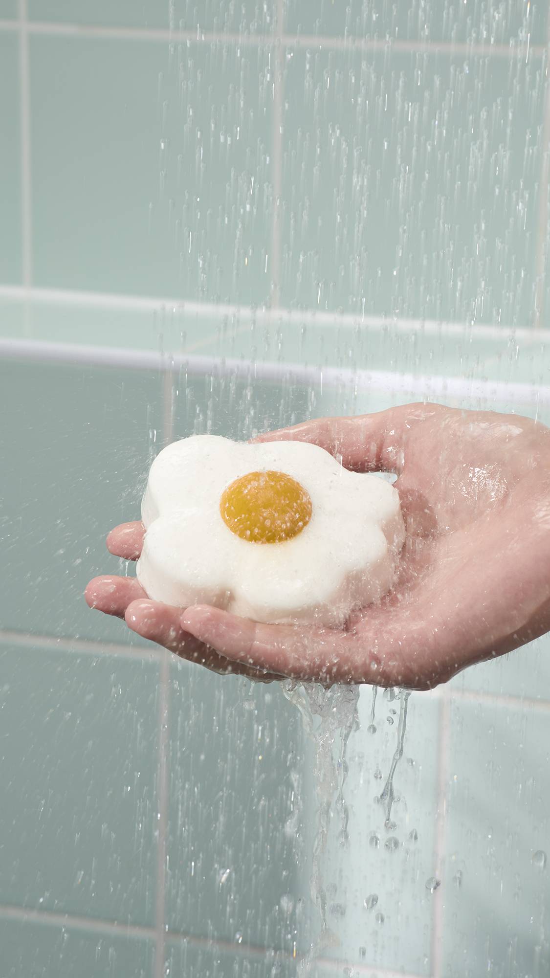 The image shows a whimsical, fried egg-shaped "Sunny Side Up" shower jelly with a bright yellow centre held in a model's open palm. The product is being rinsed under falling water in a shower against a backdrop of light blue tiles.