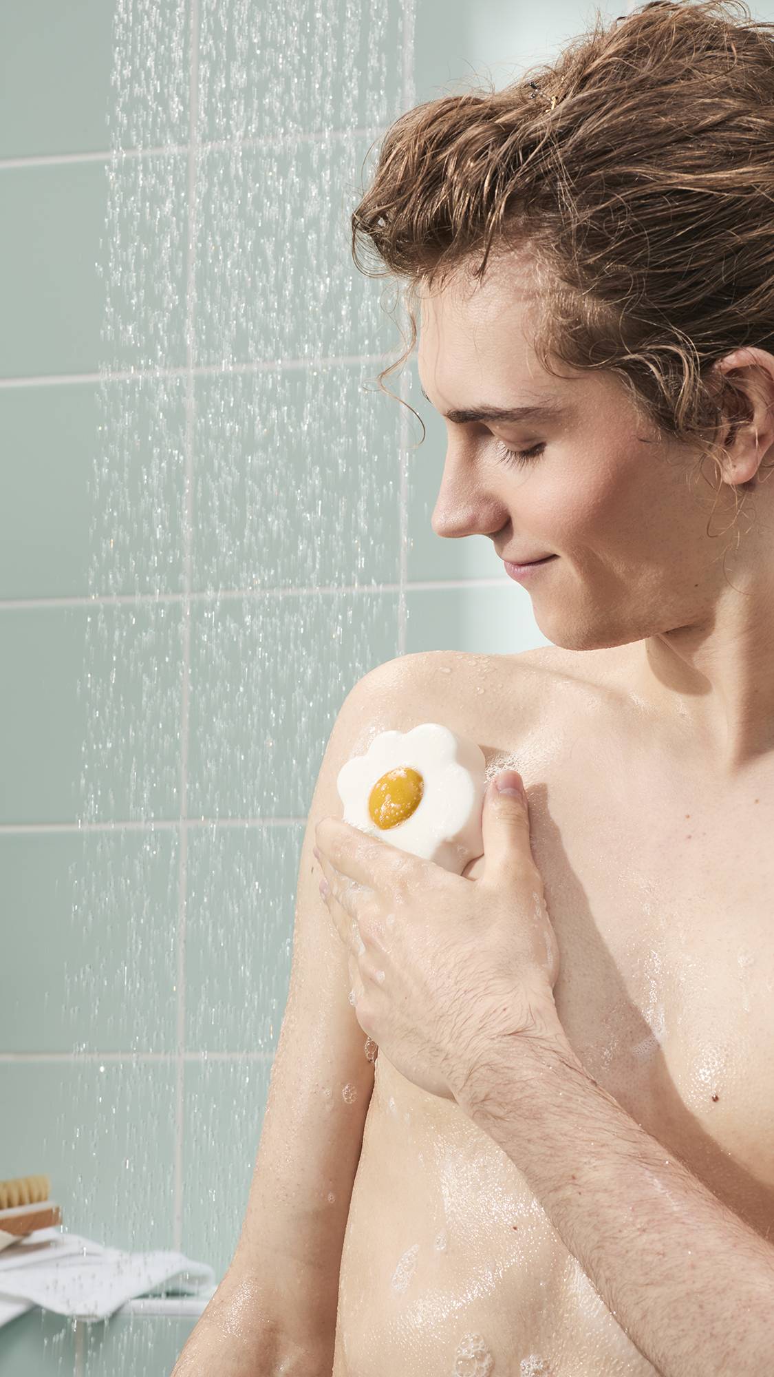 The image shows a model with damp hair smiling while using the "Sunny Side Up" fried egg-shaped shower jelly on their shoulder. They are in a shower with falling water and light blue wall tiles. 