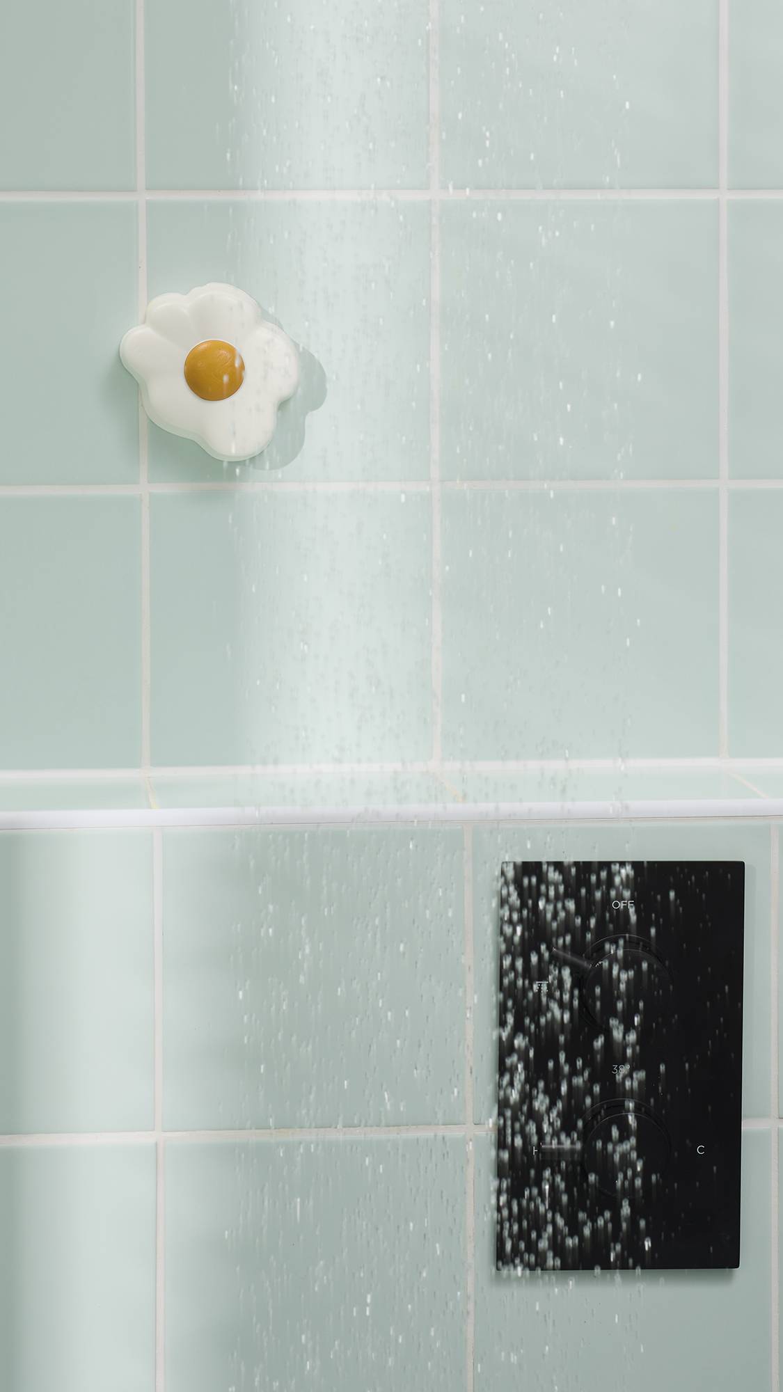 The image shows a fried egg-shaped shower jelly stuck to light green wall tiles in a shower. Water falls in the foreground, and a black digital shower control panel is visible in the bottom right corner.