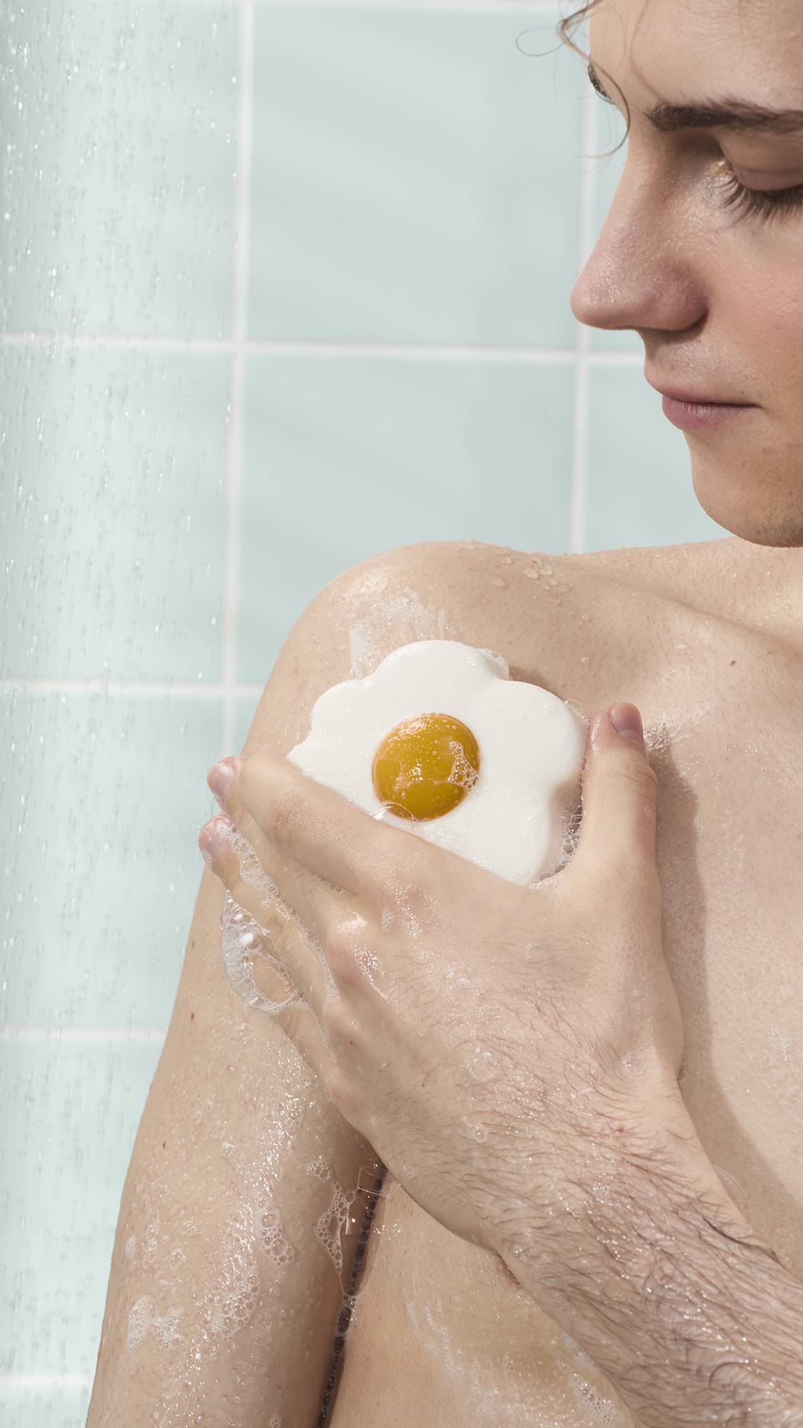 The image shows a model using a white and yellow fried egg-shaped shower jelly product to create a soapy lather on their skin. They are holding the soapy product against their shoulder, with water falling in the background against pale blue tiles.