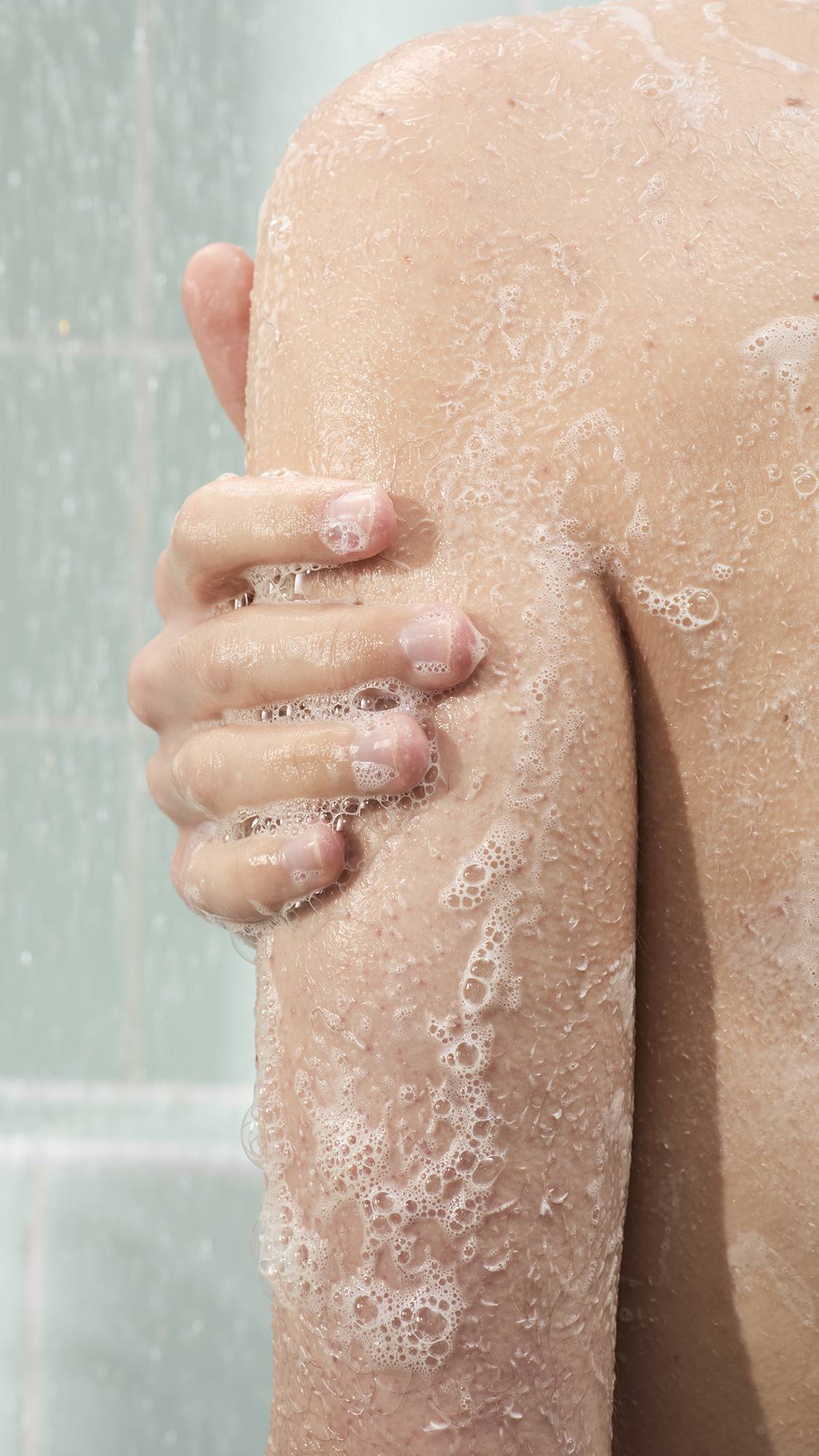 A close-up shot of a person's arm and shoulder covered in white soap bubbles in the shower. A hand is shown rubbing the lather into the skin against a background of pale green wall tiles and falling water.