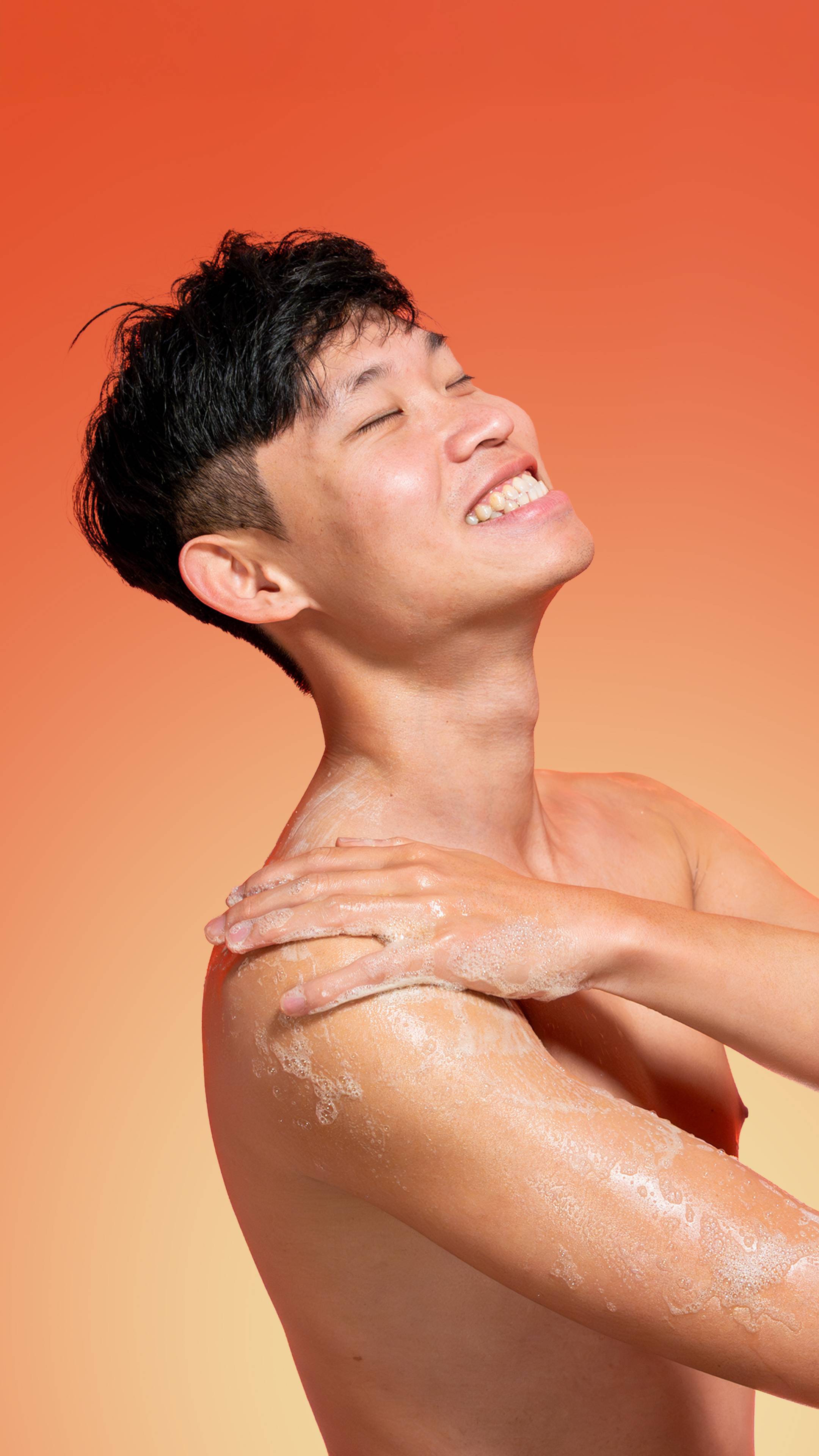 A smiling person with dark hair washes their shoulder with the sudsy lather of Sunrise shower Gel against a warm orange background.