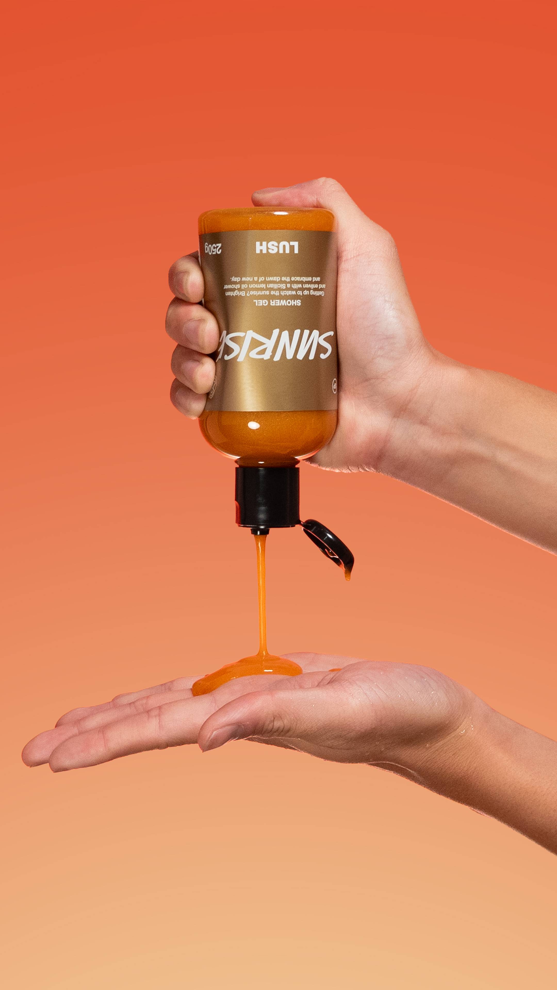 A person pours shimmering orange Sunrise shower Gel containing Plastic Free Glitter from a bottle into their open palm against a warm orange background.