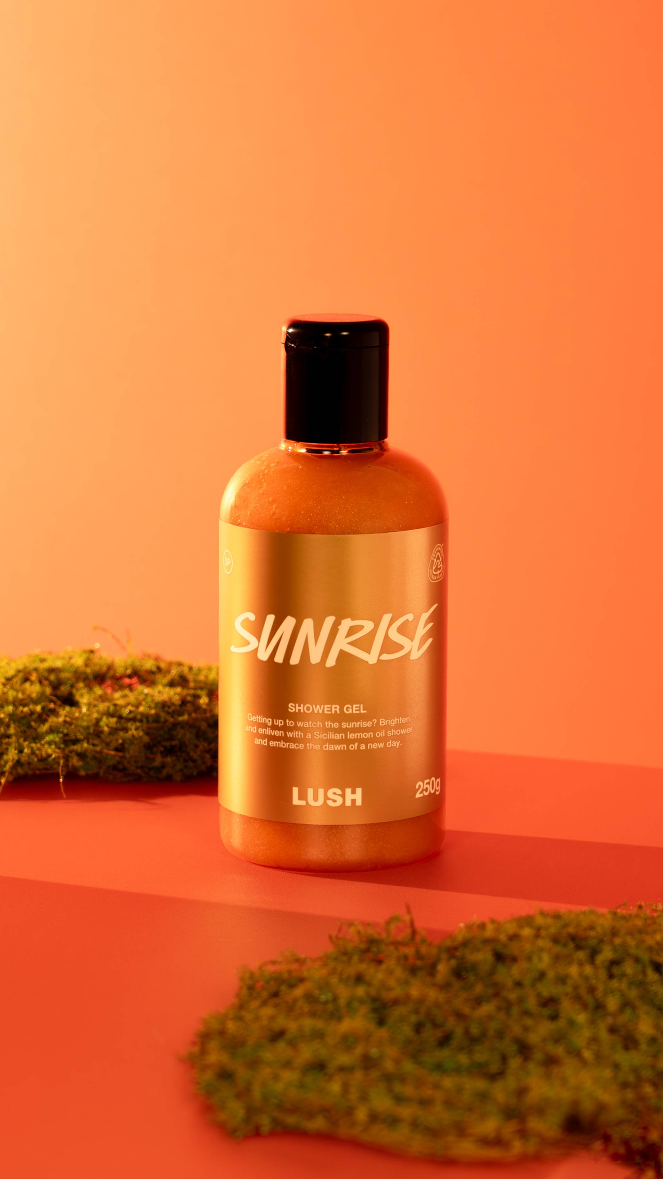 A bottle of Sunrise shower Gel filled with orange liquid and Plastic Free Glitter sits on a red surface surrounded by patches of green moss against a warm orange background.