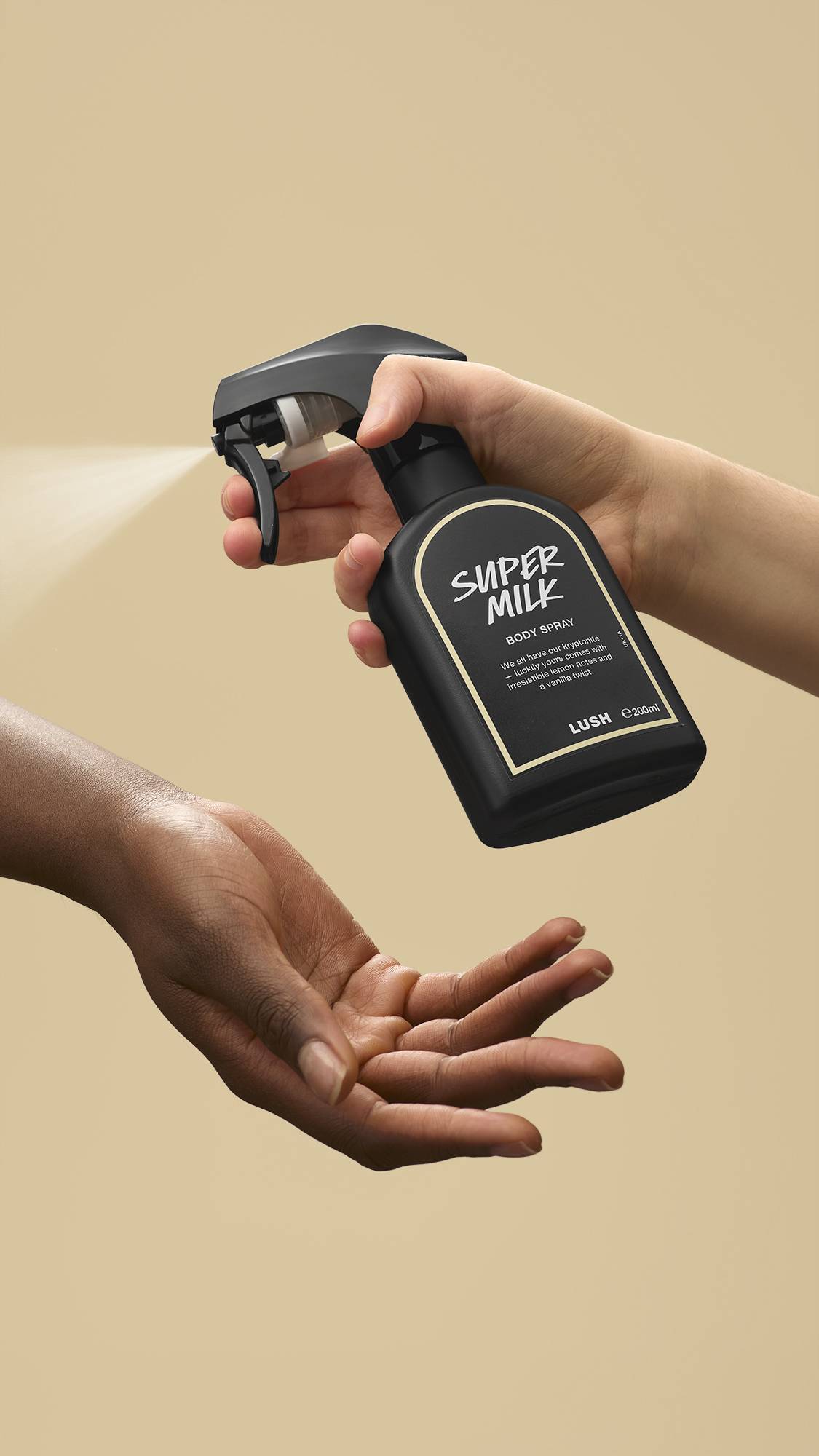 The image shows a light-toned hand spraying a bottle of LUSH 'Super Milk' body spray. A warm-toned hand is cupped below, catching the fine mist against a warm, beige background.