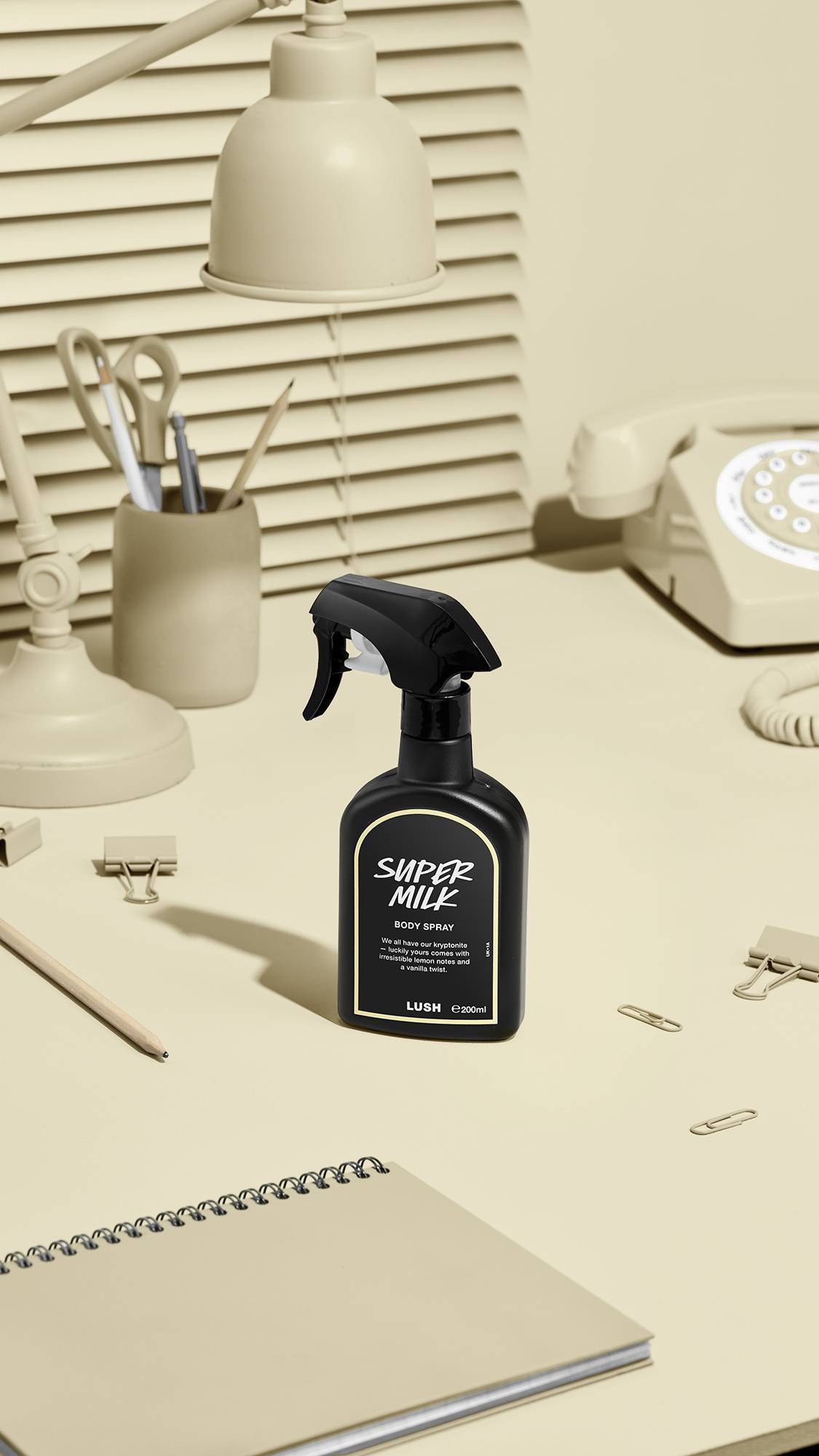 The image shows a black LUSH 'Super Milk' body spray bottle with a pale beige label on a beige desk. Nearby sit a matching desk lamp, a rotary phone, stationery, and a notebook, against window blinds.