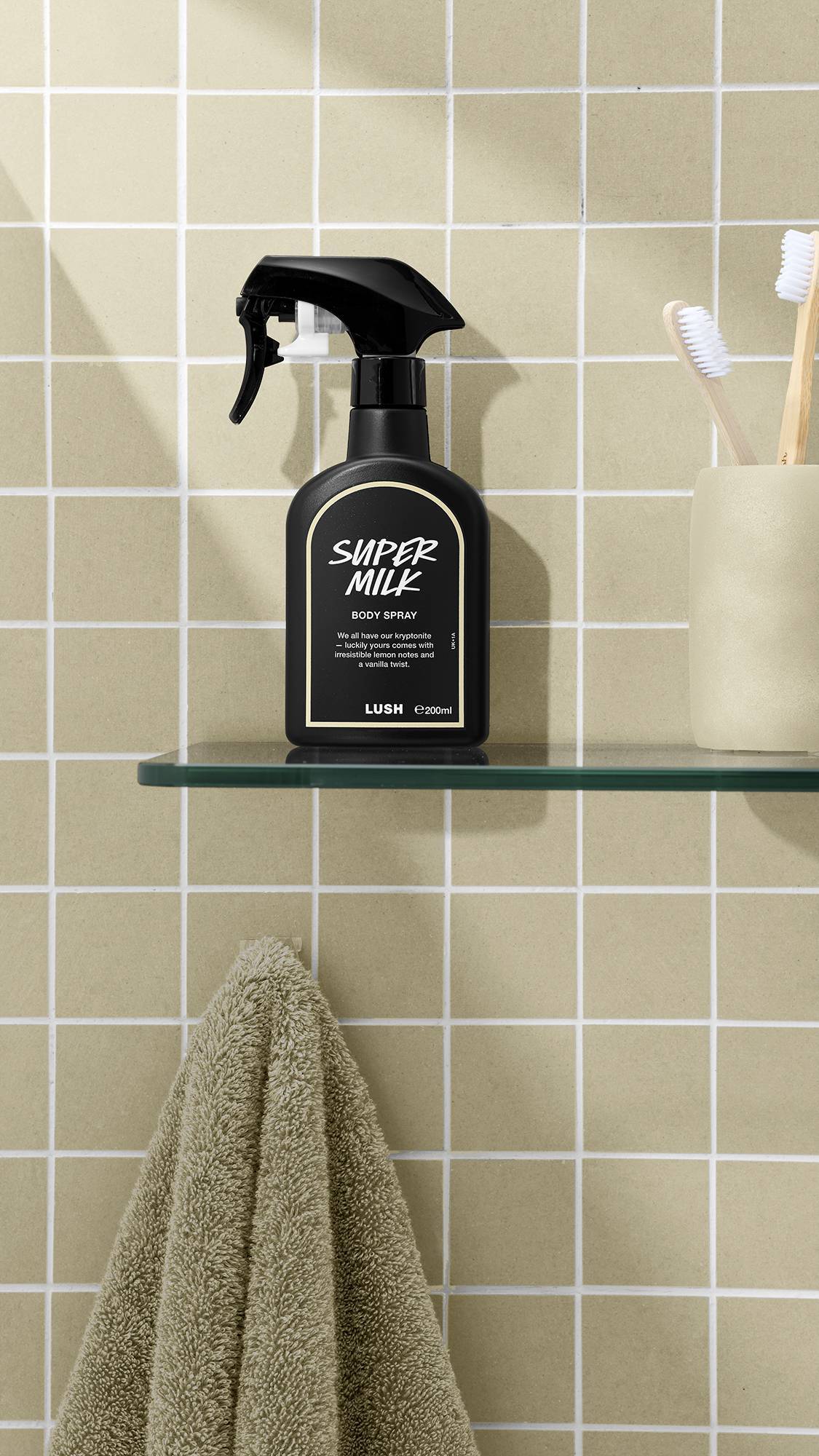 The image shows a bottle of LUSH 'Super Milk' body spray on a glass shelf. A beige towel hangs below, with a matching coloured cup and brushes nearby, against a warm, creamy-beige, tiled bathroom wall.