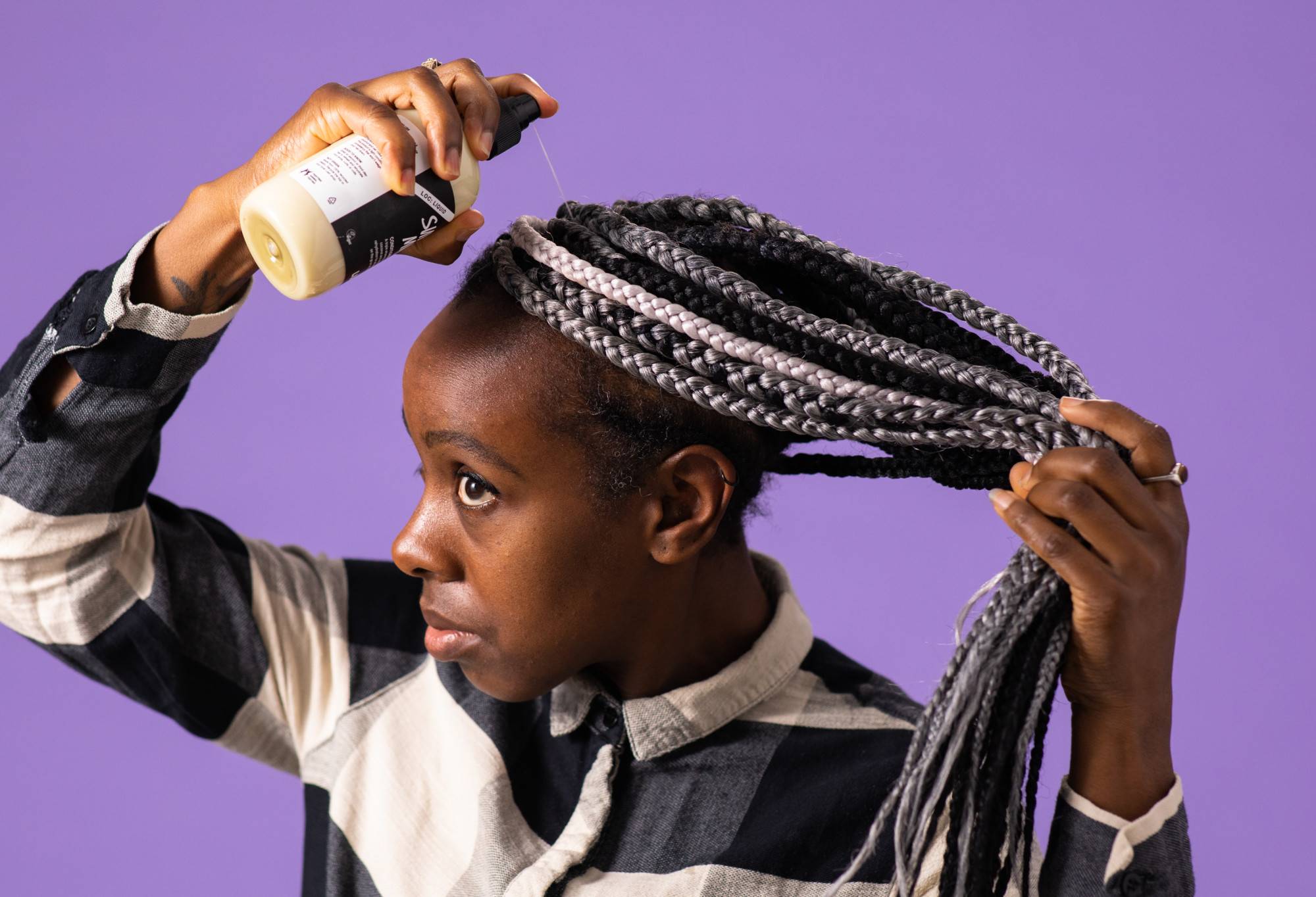 A person with long silver, grey and black braids sprays Super Milk, a light, white conditioning spray, into their roots.